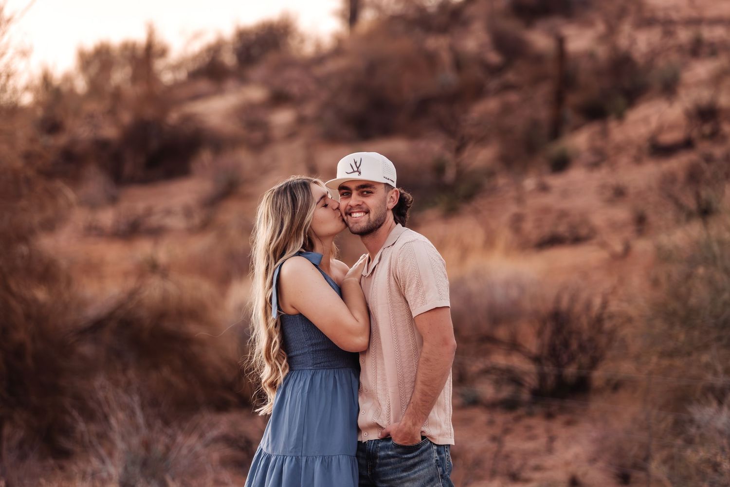 Couple embracing in warm desert landscape during golden hour with woman in blue dress and man in baseball cap.