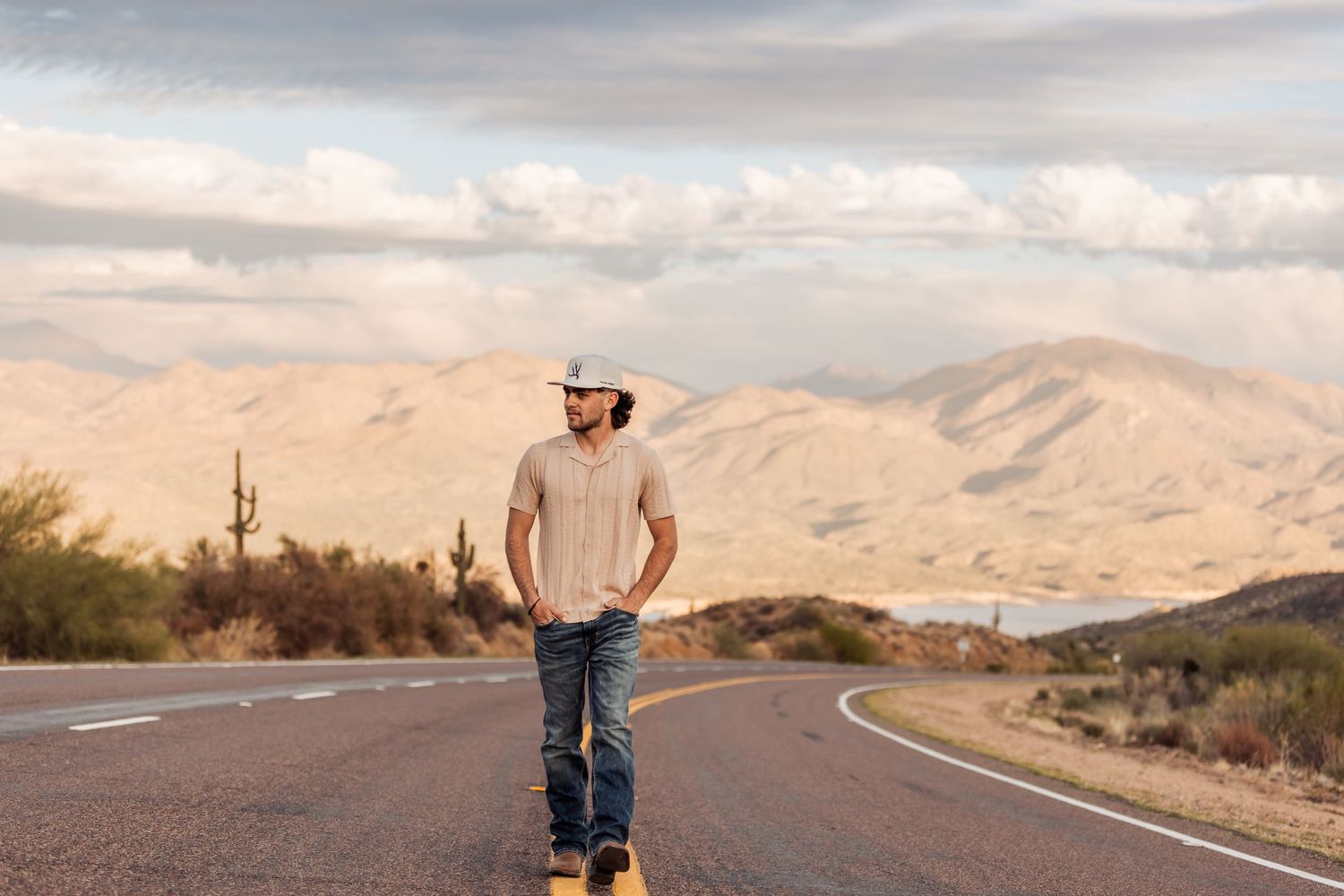 Someone in casual attire stands on a winding desert road with mountains and cloudy skies in the background.