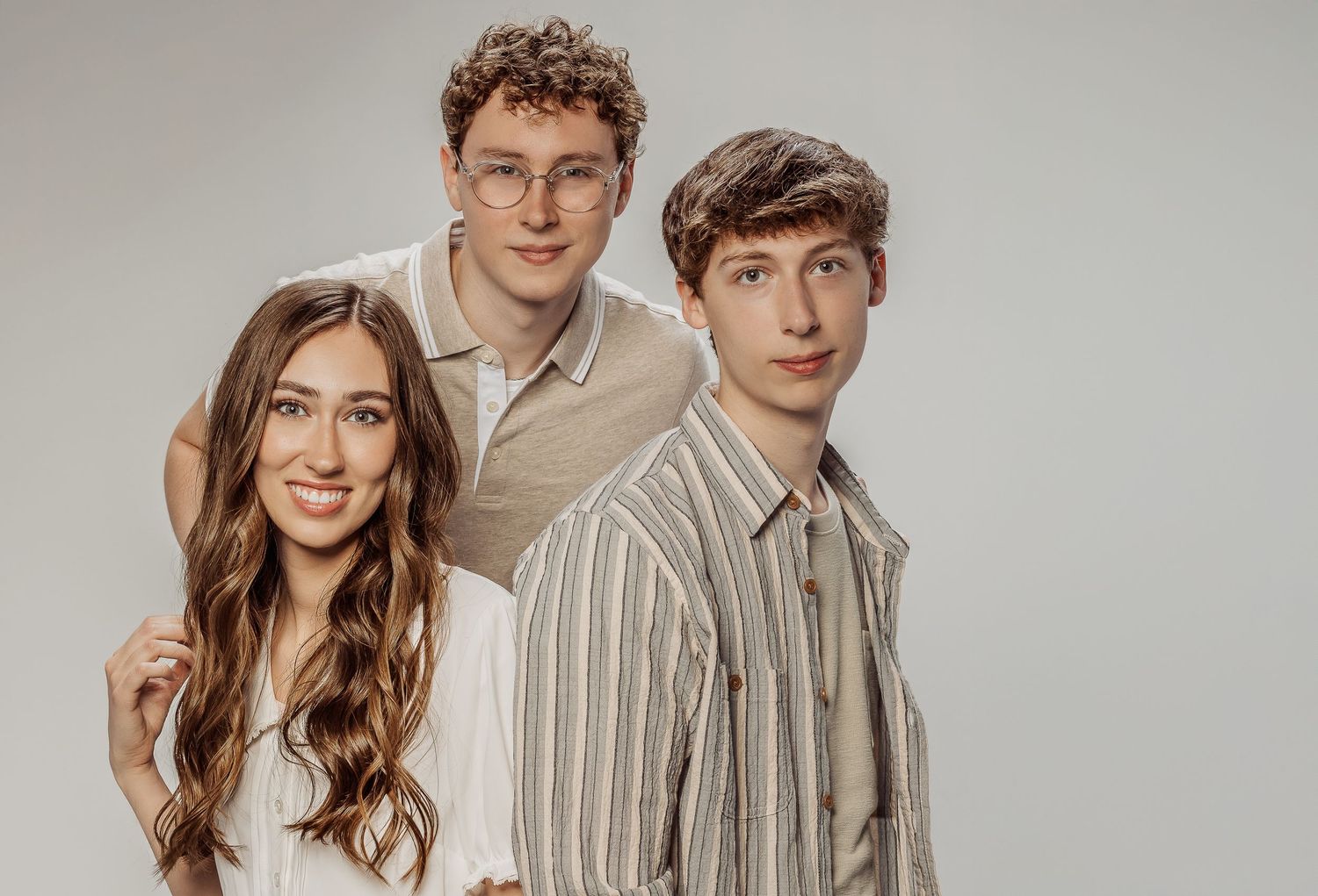 Three young people pose together against a neutral background wearing casual beige and striped button-up shirts.