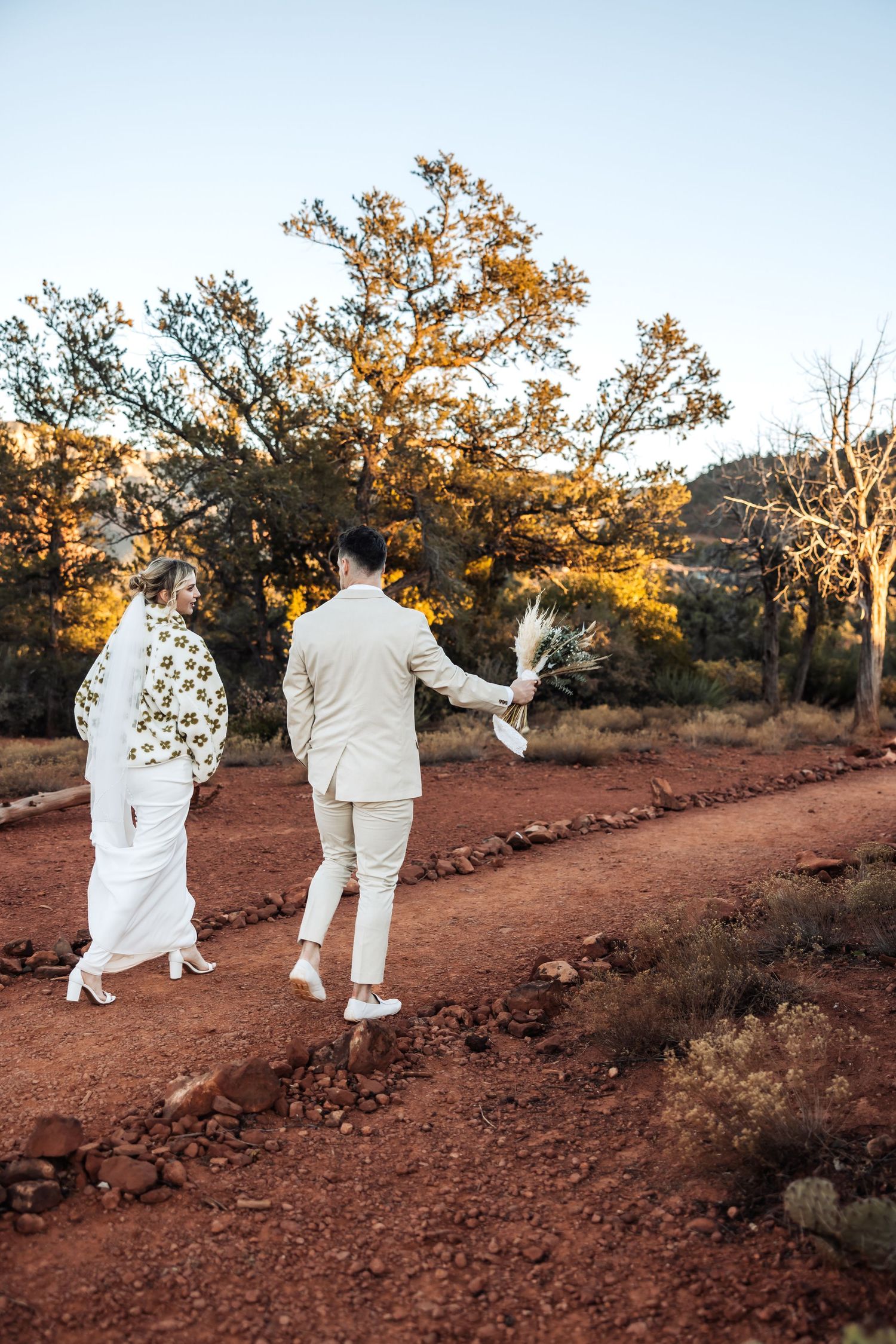 Two people in white attire walk hand in hand along a red dirt path surrounded by desert landscape at golden hour.