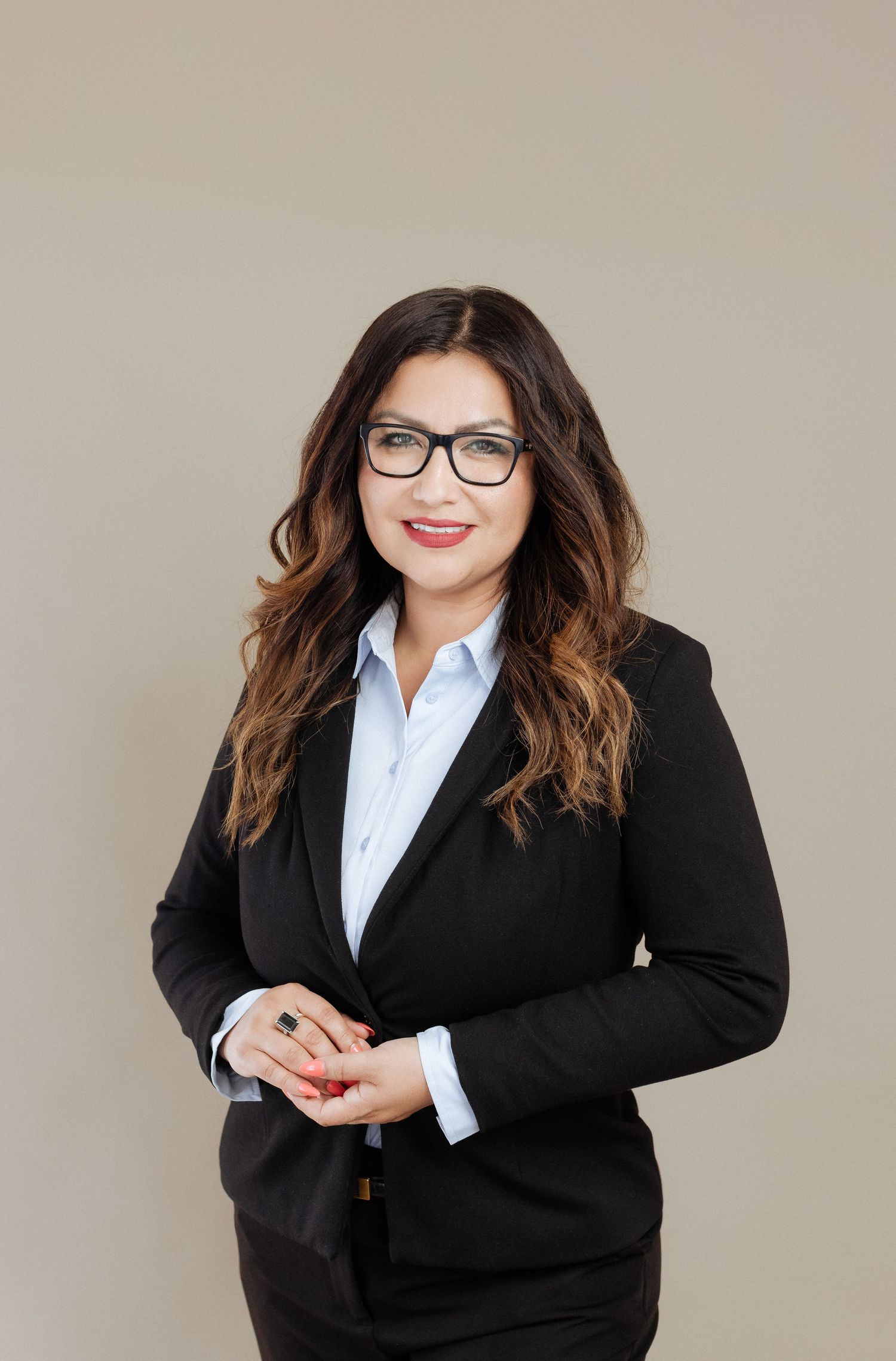 Professional wearing a black blazer suit jacket with glasses and a light blue button-down shirt against neutral backdrop.