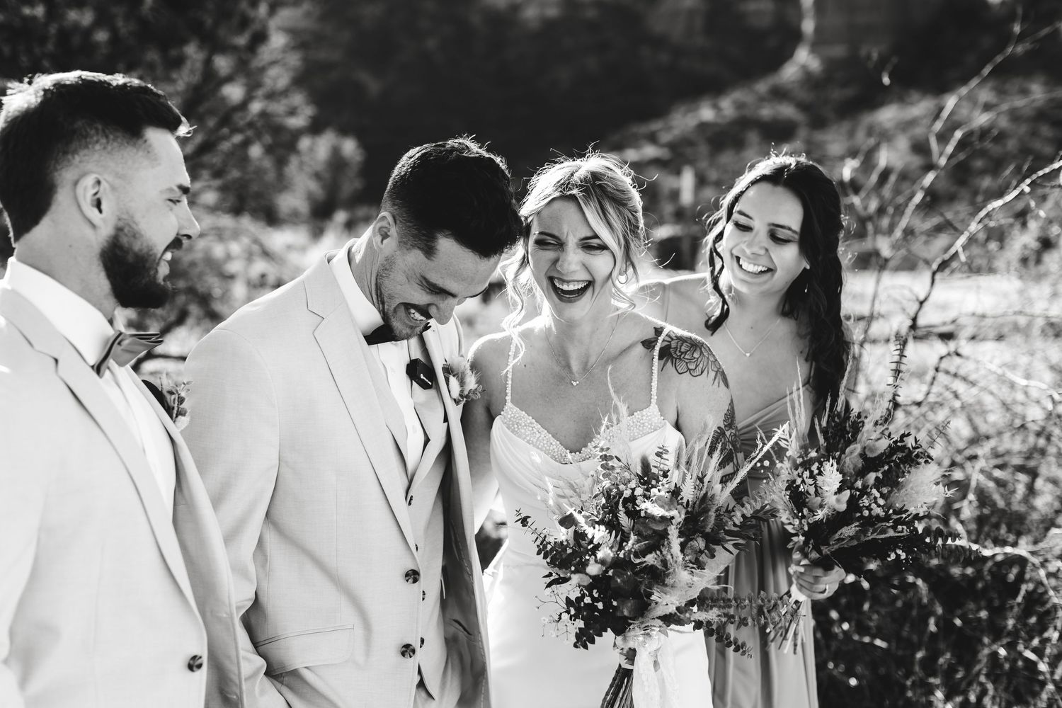 Black and white photo of a wedding party laughing together outdoors while holding floral bouquets.