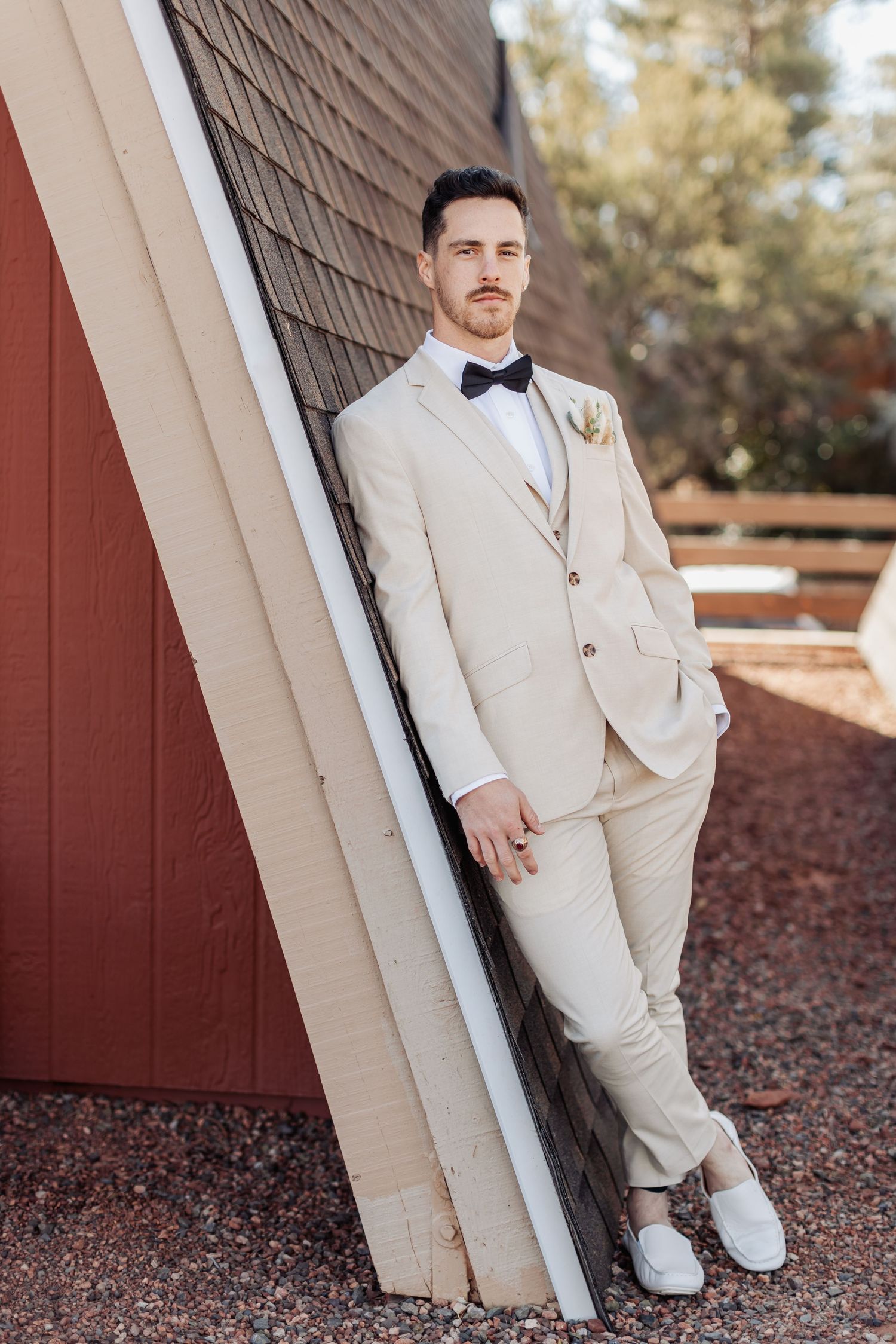 Close-up of a groom in a light beige wedding suit and black bow tie leaning against a white pillar outdoors.