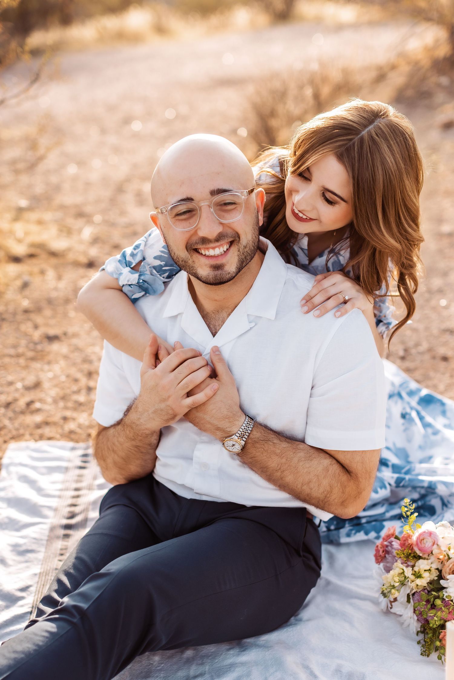 Couple sharing an intimate moment while sitting on a blanket during sunset with flowers nearby.