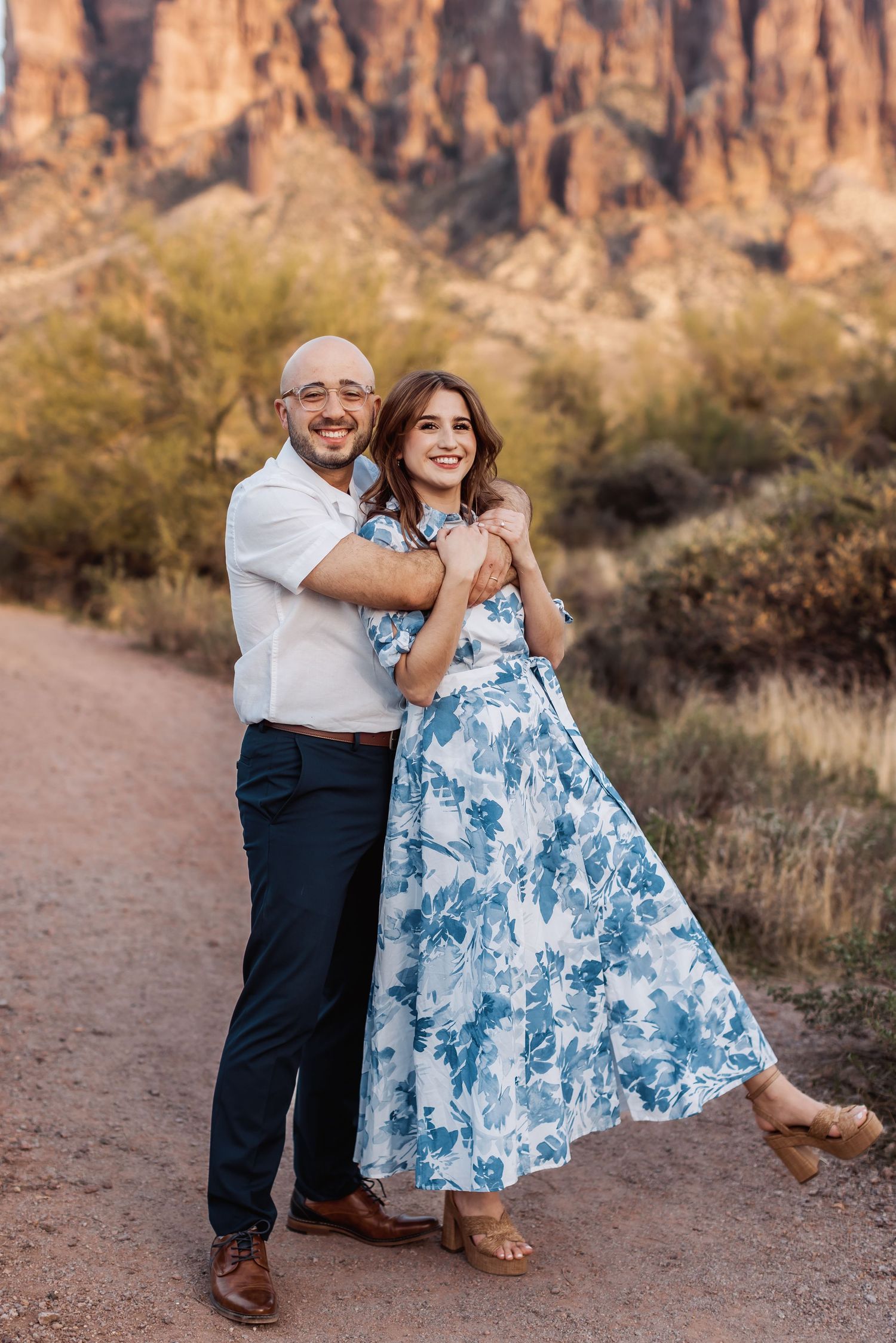 A joyful couple embraces on a desert trail with towering red rock cliffs and desert brush in the background.