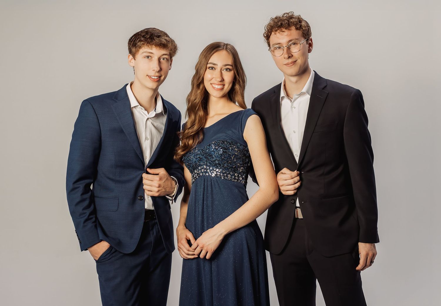 Three elegantly dressed people pose together in front of a light gray backdrop in formal attire.