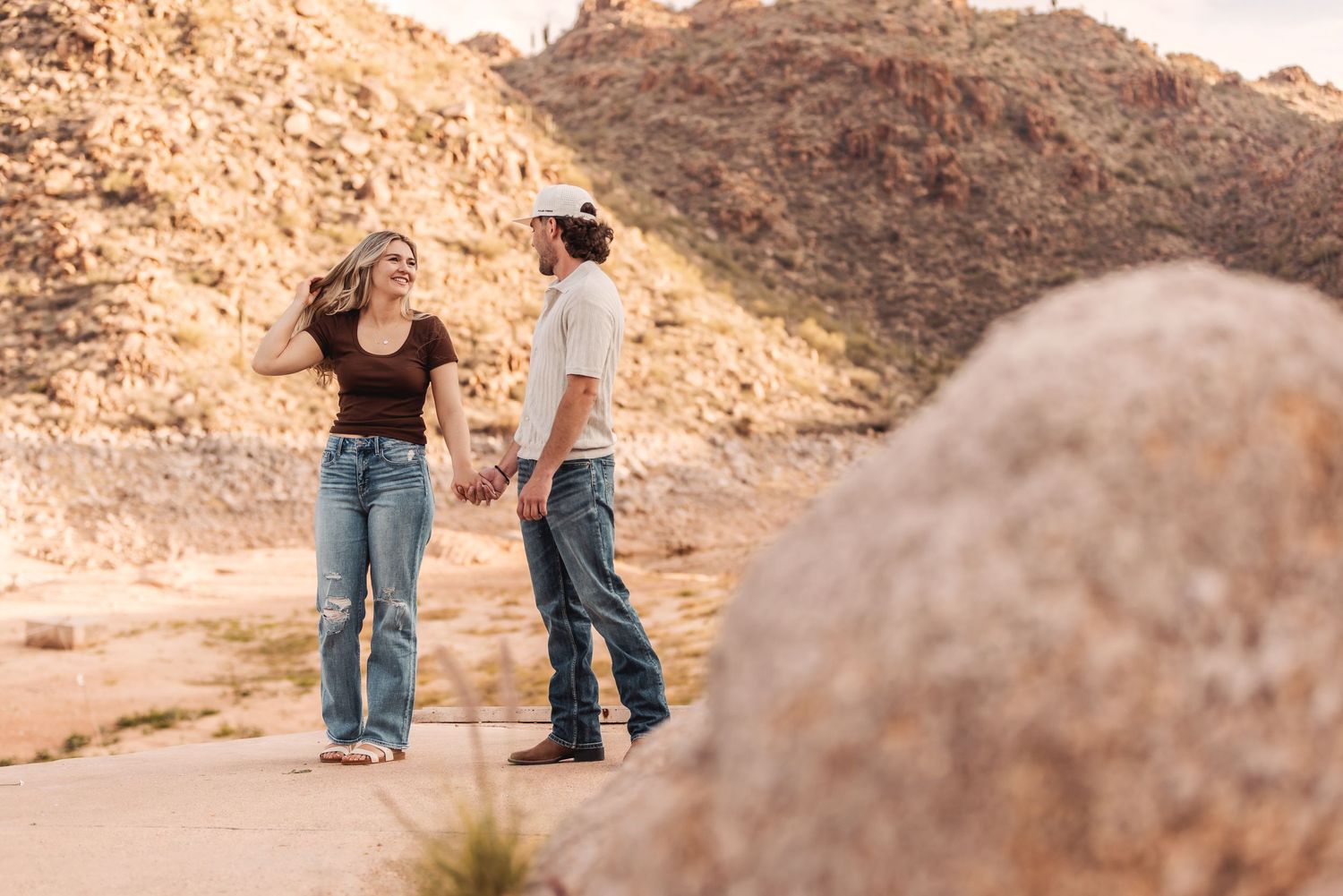Two people hold hands while walking along a desert trail surrounded by rugged rocky mountains in warm sunlight.