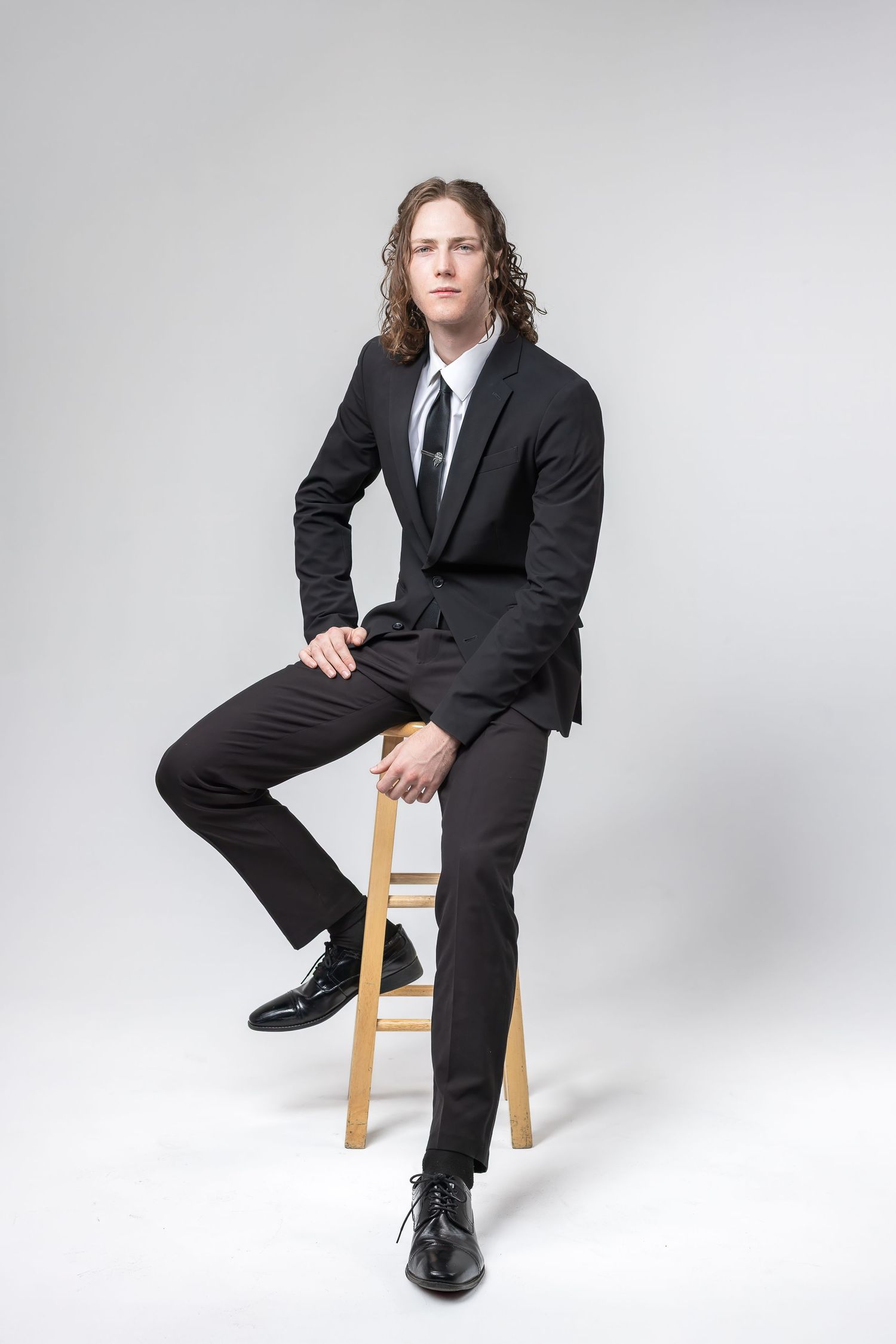 Professional studio portrait of someone with long curly hair in a black suit and tie sitting on a wooden stool.