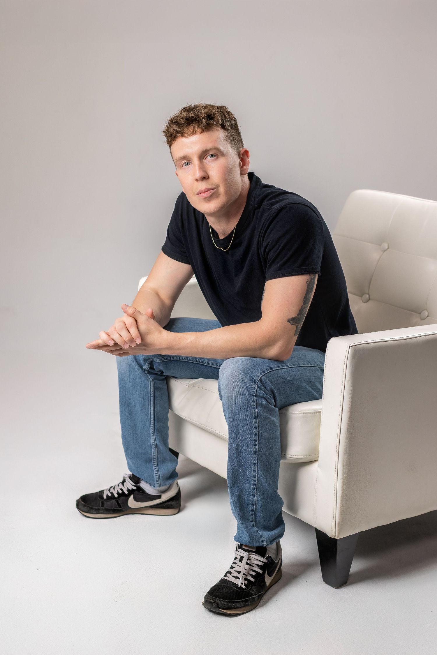 A person in jeans and a black shirt sits on a white chair in various casual poses against a light background.