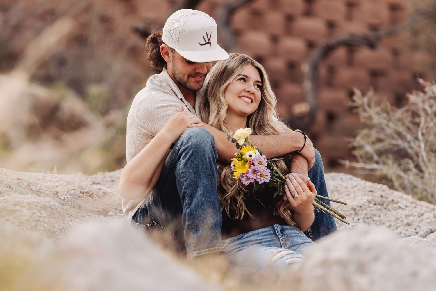 A couple sits together on a rocky outcrop, the woman holding a colorful bouquet of wildflowers in her hands.