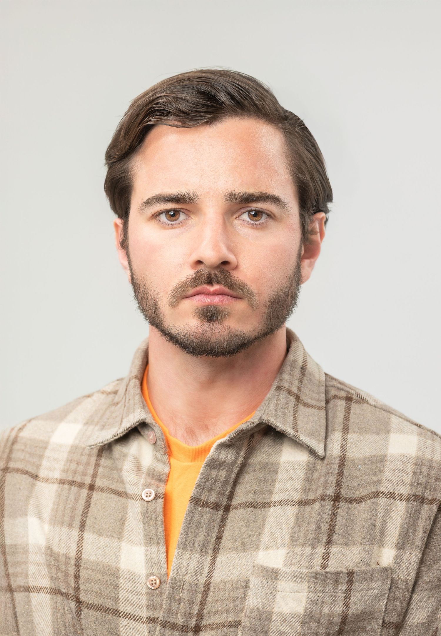 Portrait wearing plaid brown button-up shirt over orange t-shirt against light gray background.