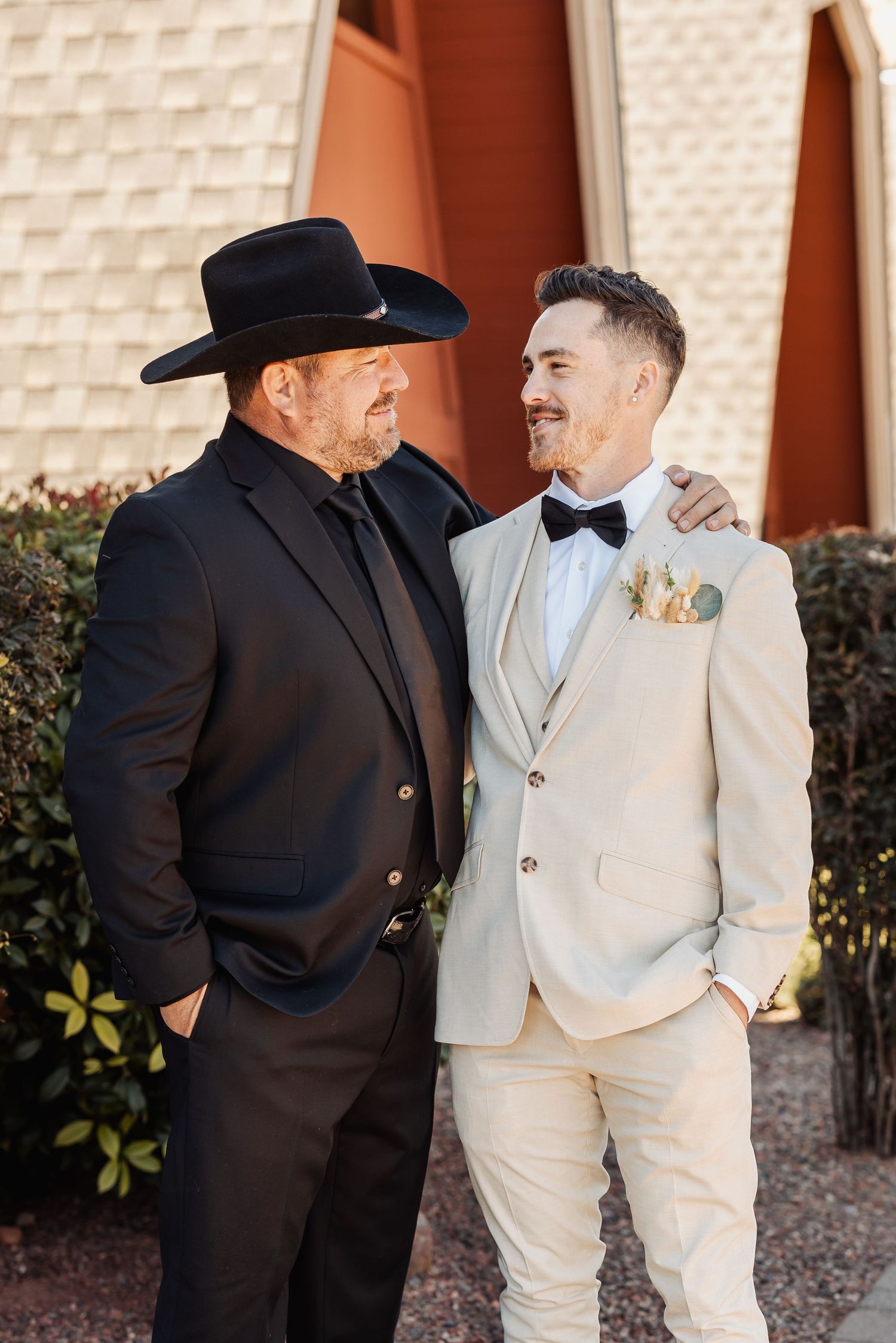 Two people in contrasting formal attire, one in black with cowboy hat and one in cream-colored suit with bowtie, embrace smiling.