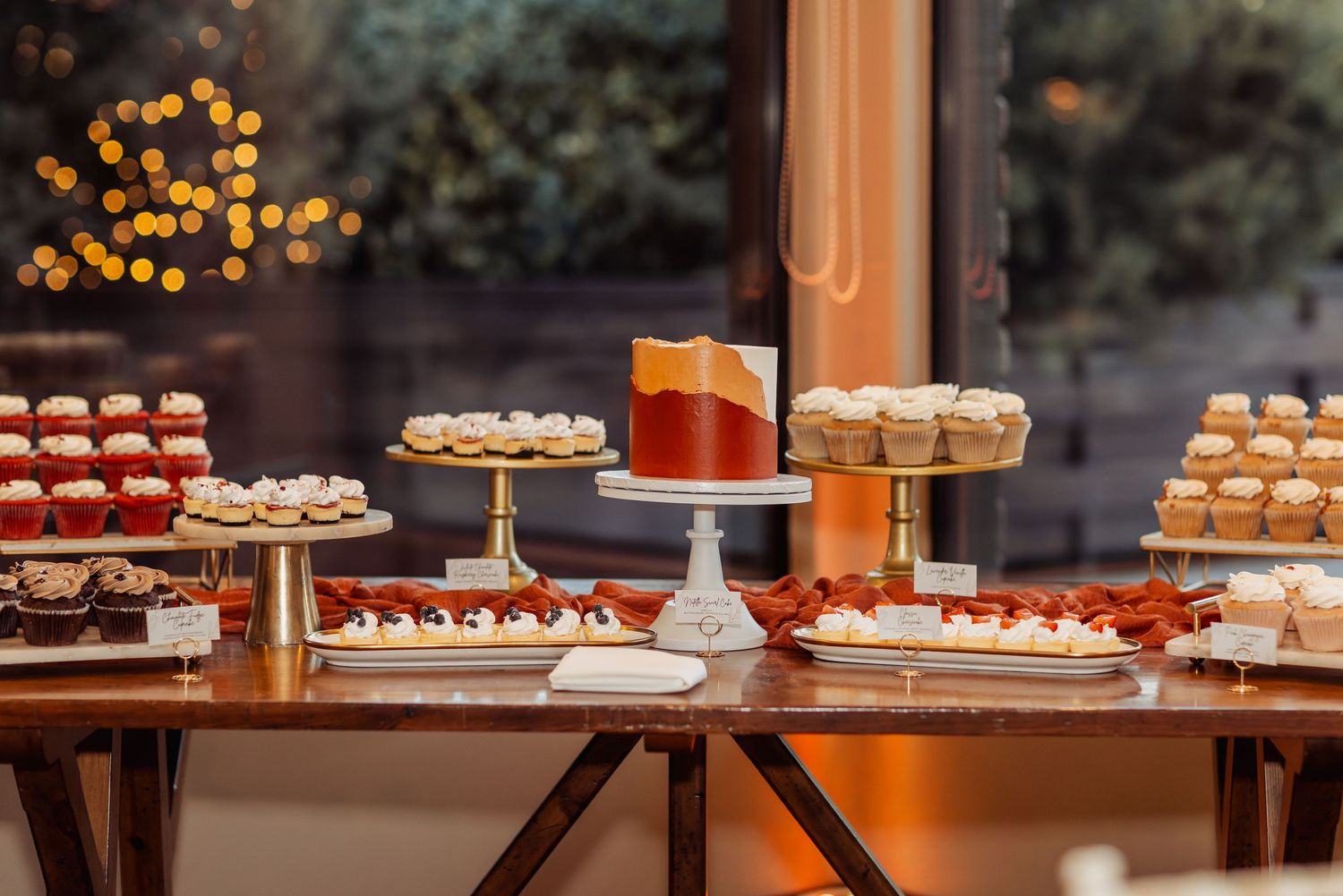 Elegant dessert display with tiered stands showcasing cupcakes and pastries in front of window with warm lighting.