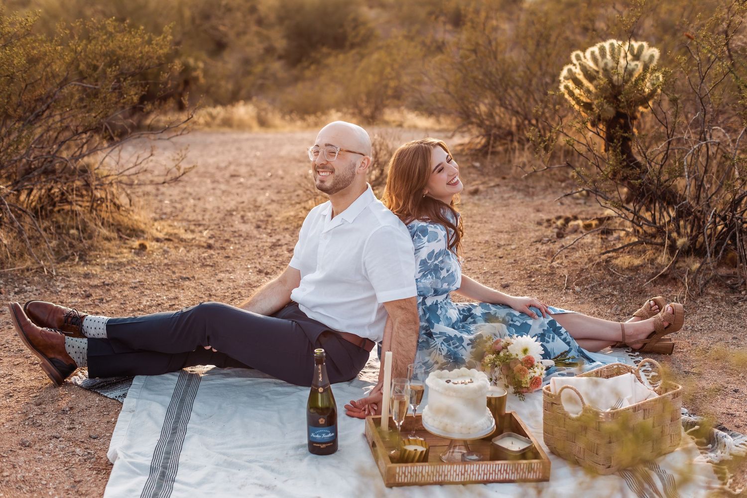 A couple enjoys a romantic picnic on a blanket in the desert with wine and snacks at golden hour.