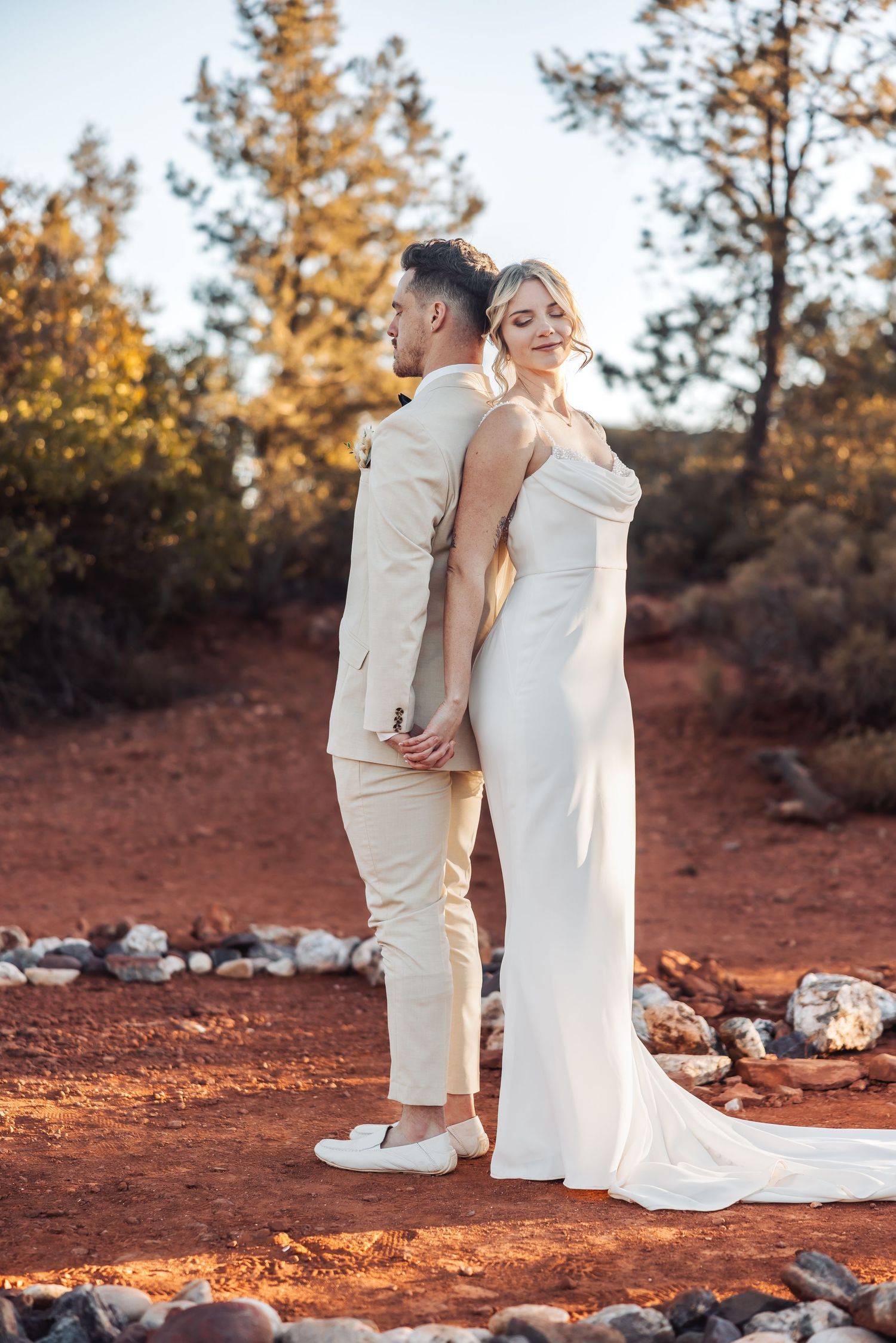 Bride and groom in all-white attire pose back-to-back on red desert terrain with pine trees in background.