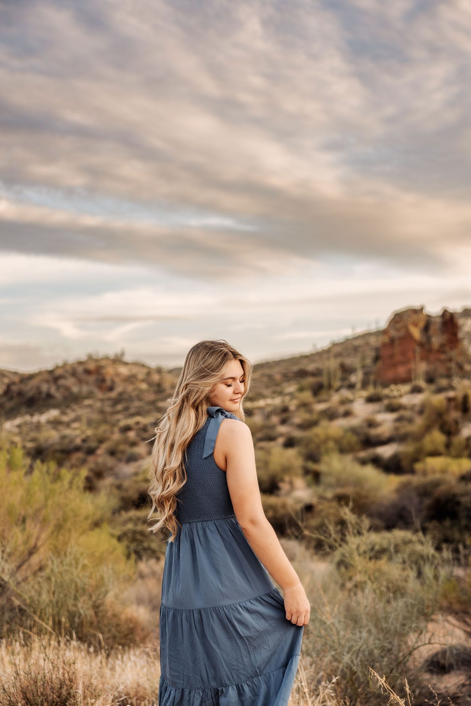 A woman in a flowing blue dress stands against a desert landscape with rocky mountains and cloudy sunset skies.