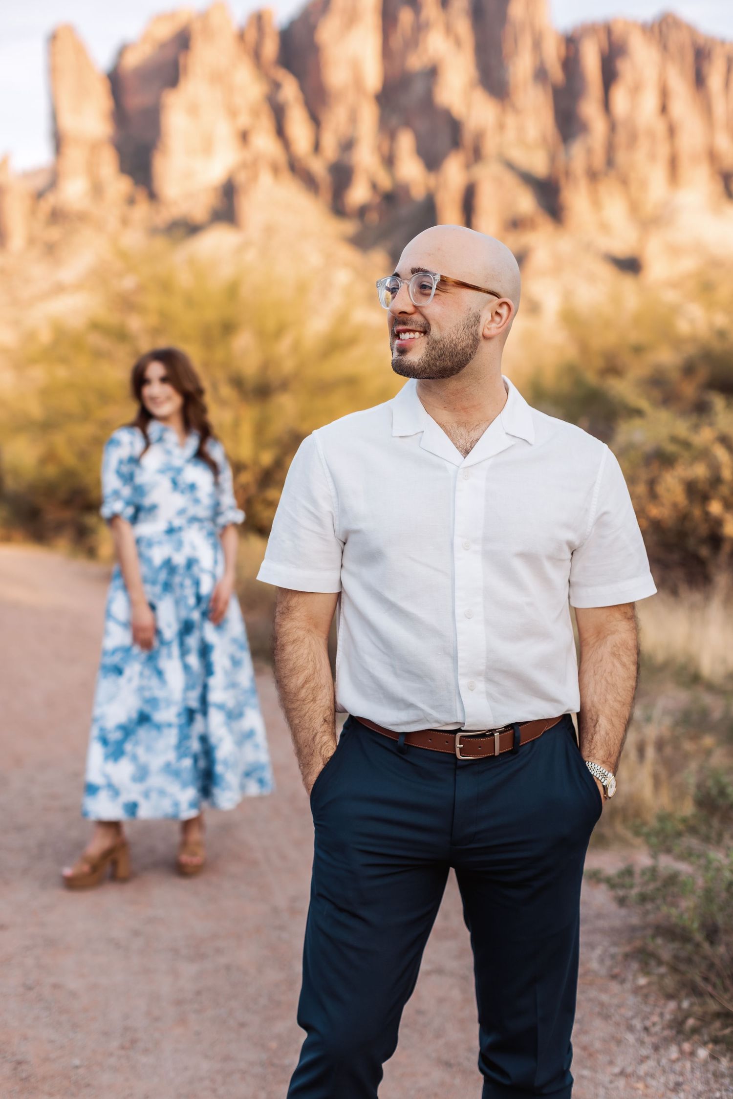 A couple poses for photos on a desert trail with majestic red rock formations in the background at sunset.