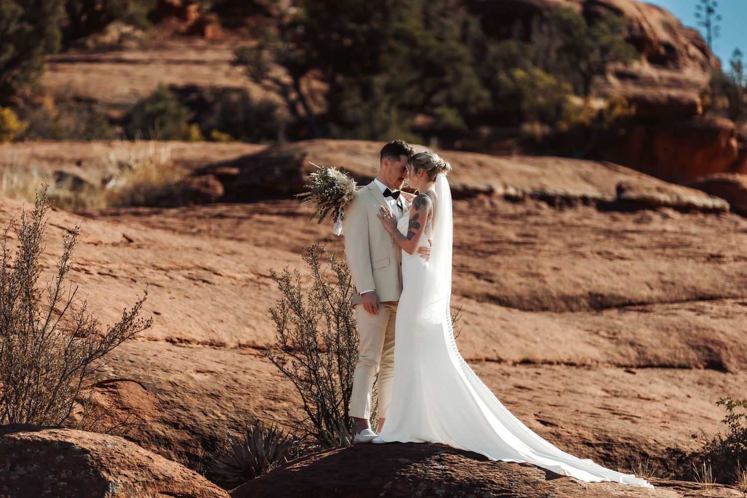 A bride in a flowing white wedding dress poses gracefully against red rock formations in a desert landscape.