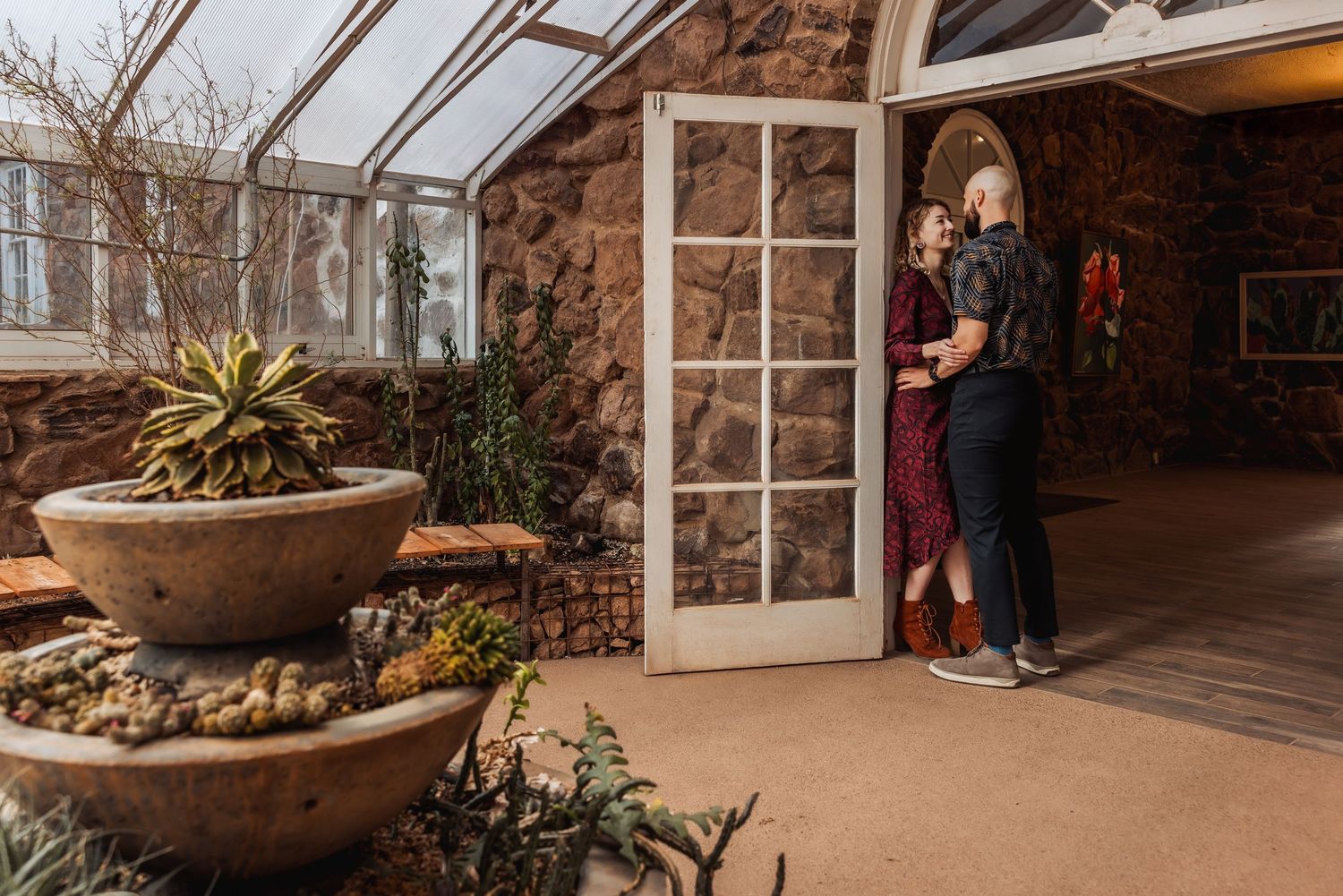 Romantic couple embraces in doorway of stone conservatory with glass ceiling and potted plants.
