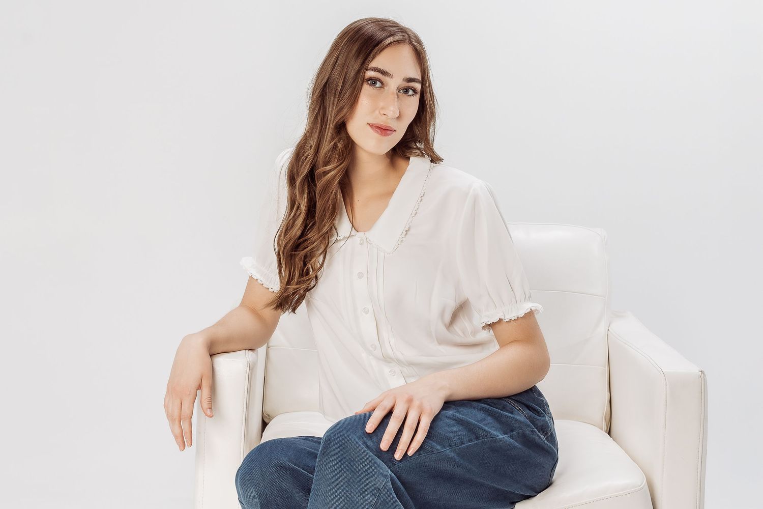 Person in light casual attire poses on white sofa in modern studio setting against white backdrop with soft lighting.