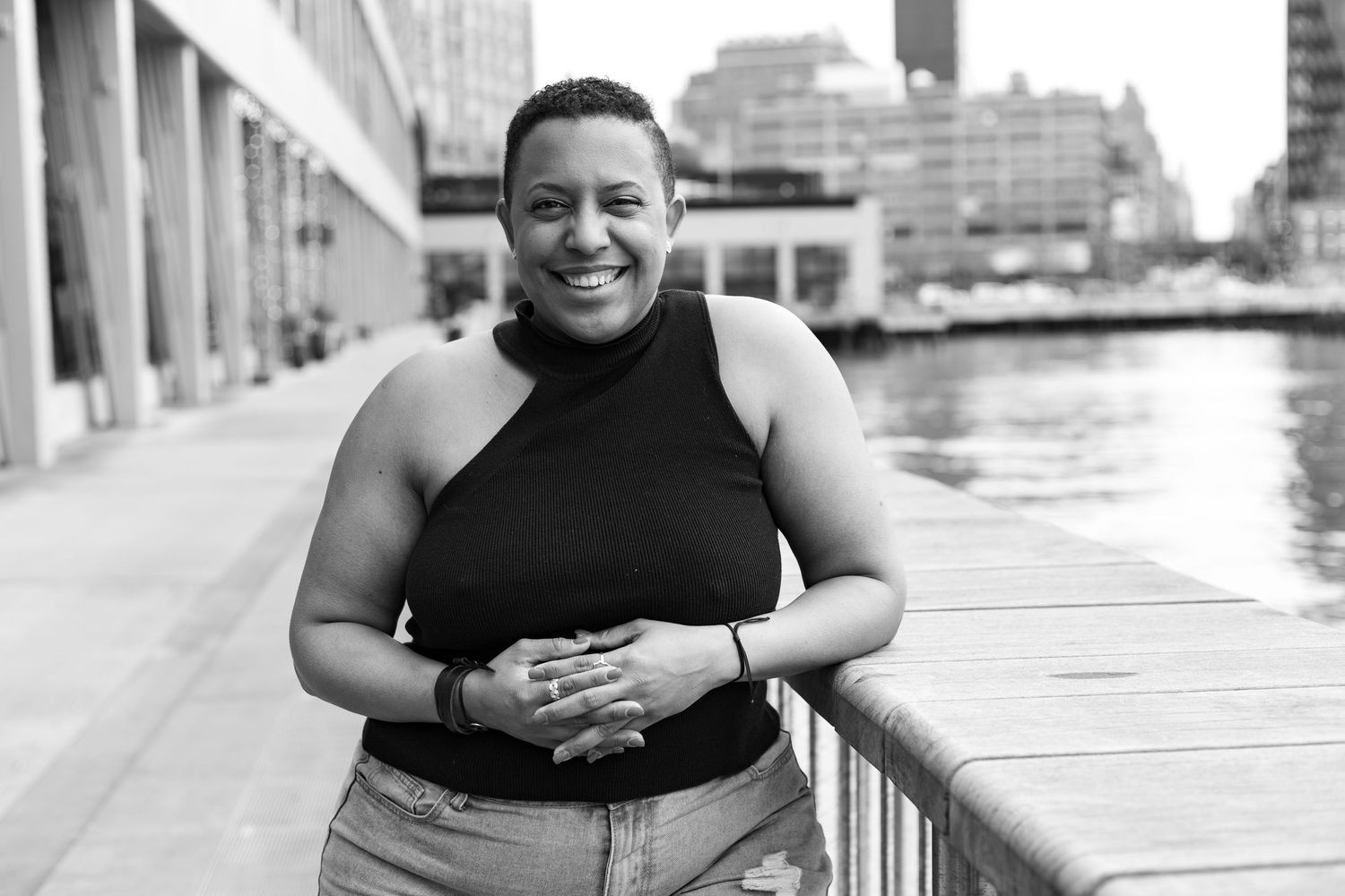 A smiling person wearing a black sleeveless top poses along a waterfront walkway in black and white photography.