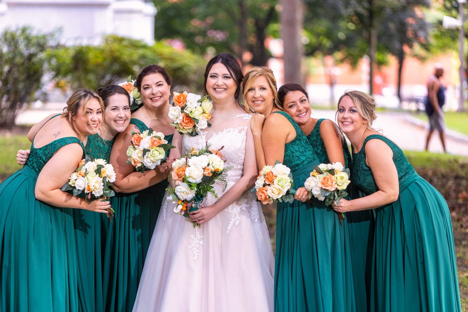 A group of bridesmaids in teal green dresses holding flower bouquets pose together outdoors for wedding photos.