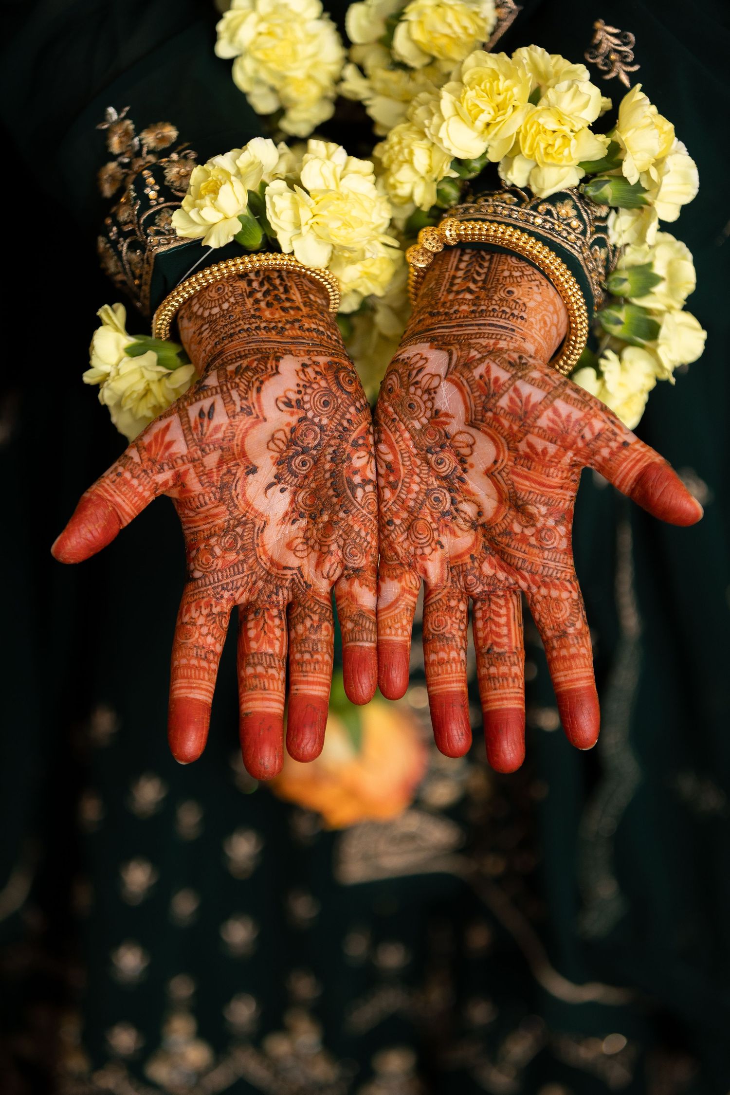 Palms with intricate henna mehndi designs displayed with yellow roses and ornate gold bangles in traditional style.