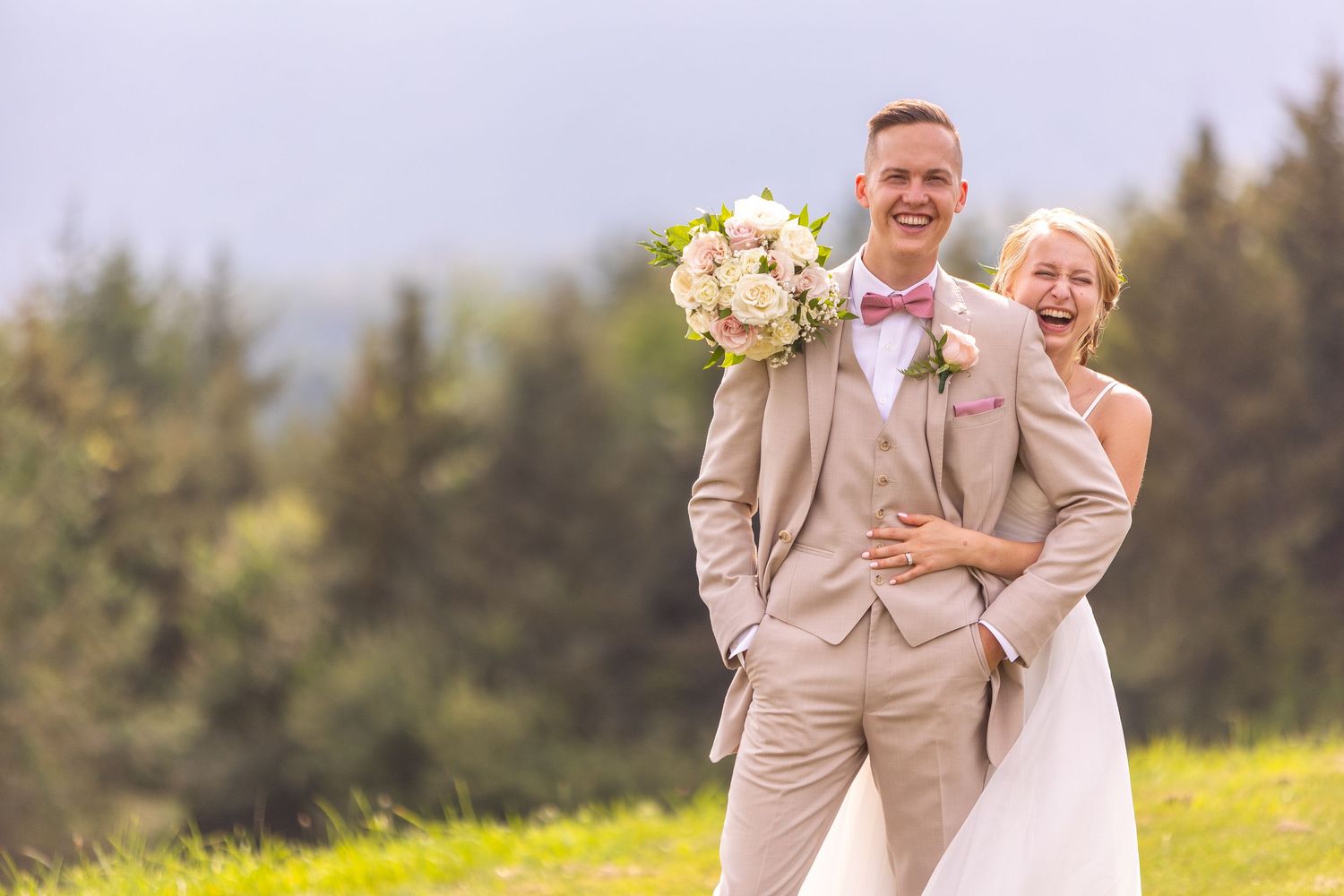 A newlywed couple in beige attire share a joyful moment on a hilltop with evergreen trees in the background.