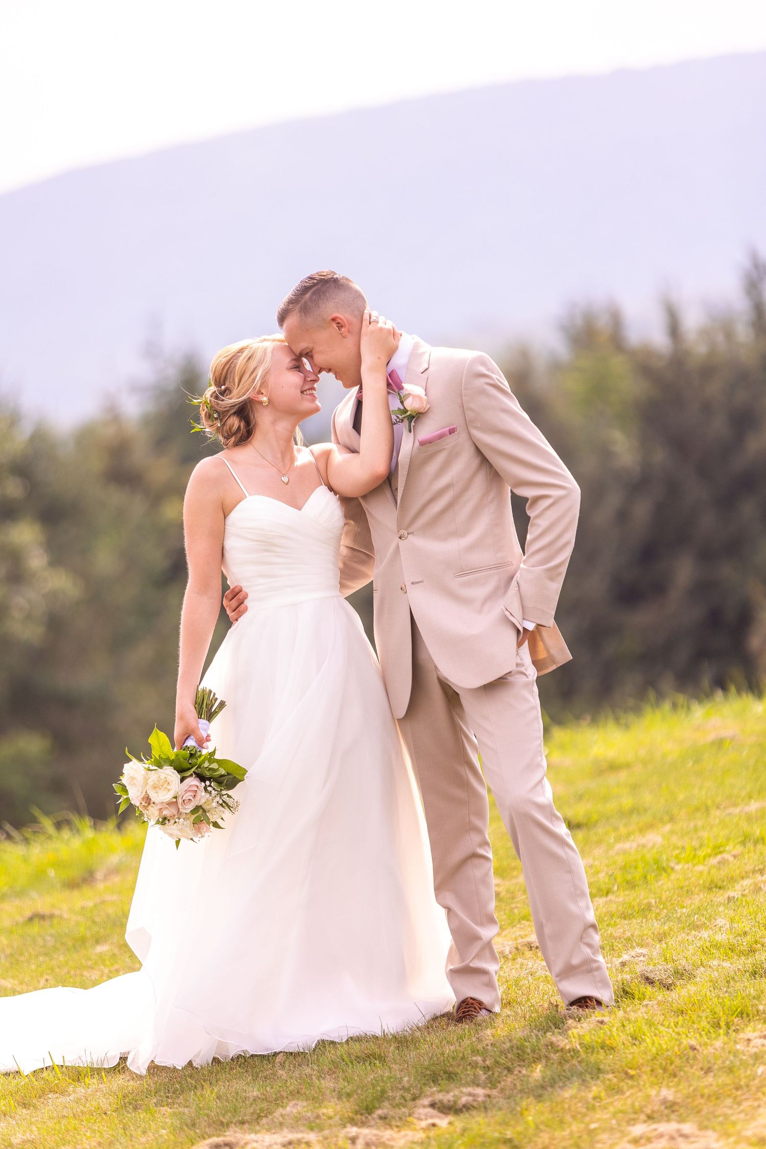 Romantic outdoor wedding portrait on a sunny hillside with a couple in tan suit and white gown holding a floral bouquet.