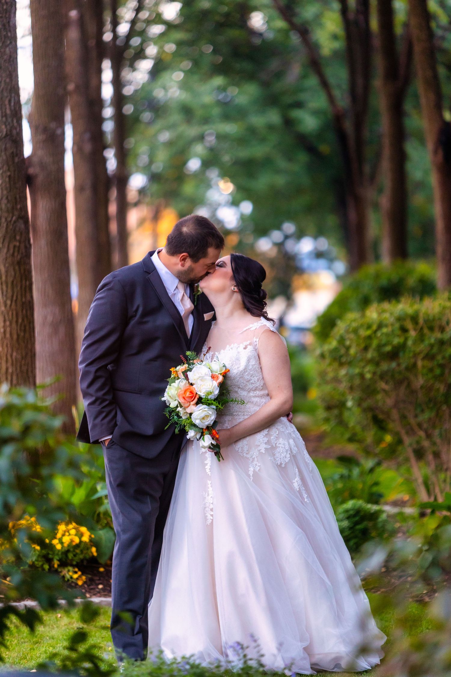 A romantic wedding scene with a couple kissing in a lush garden pathway lined with trees and yellow flowers.