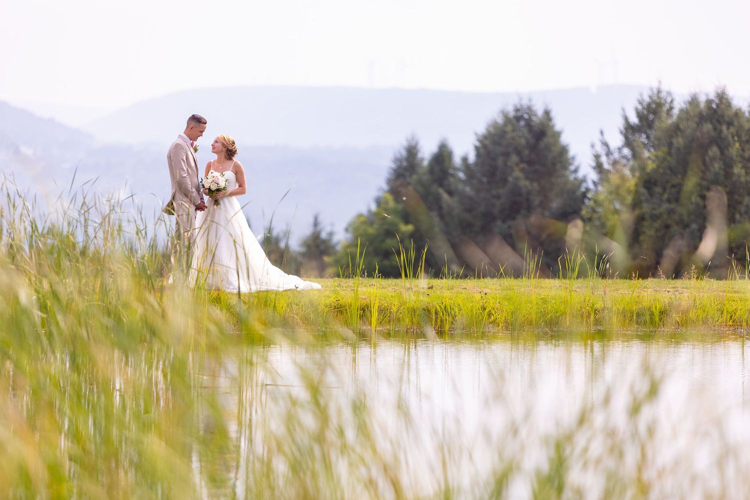 A wedding couple stands together in a scenic meadow surrounded by tall grass with mountains and forest in the background.