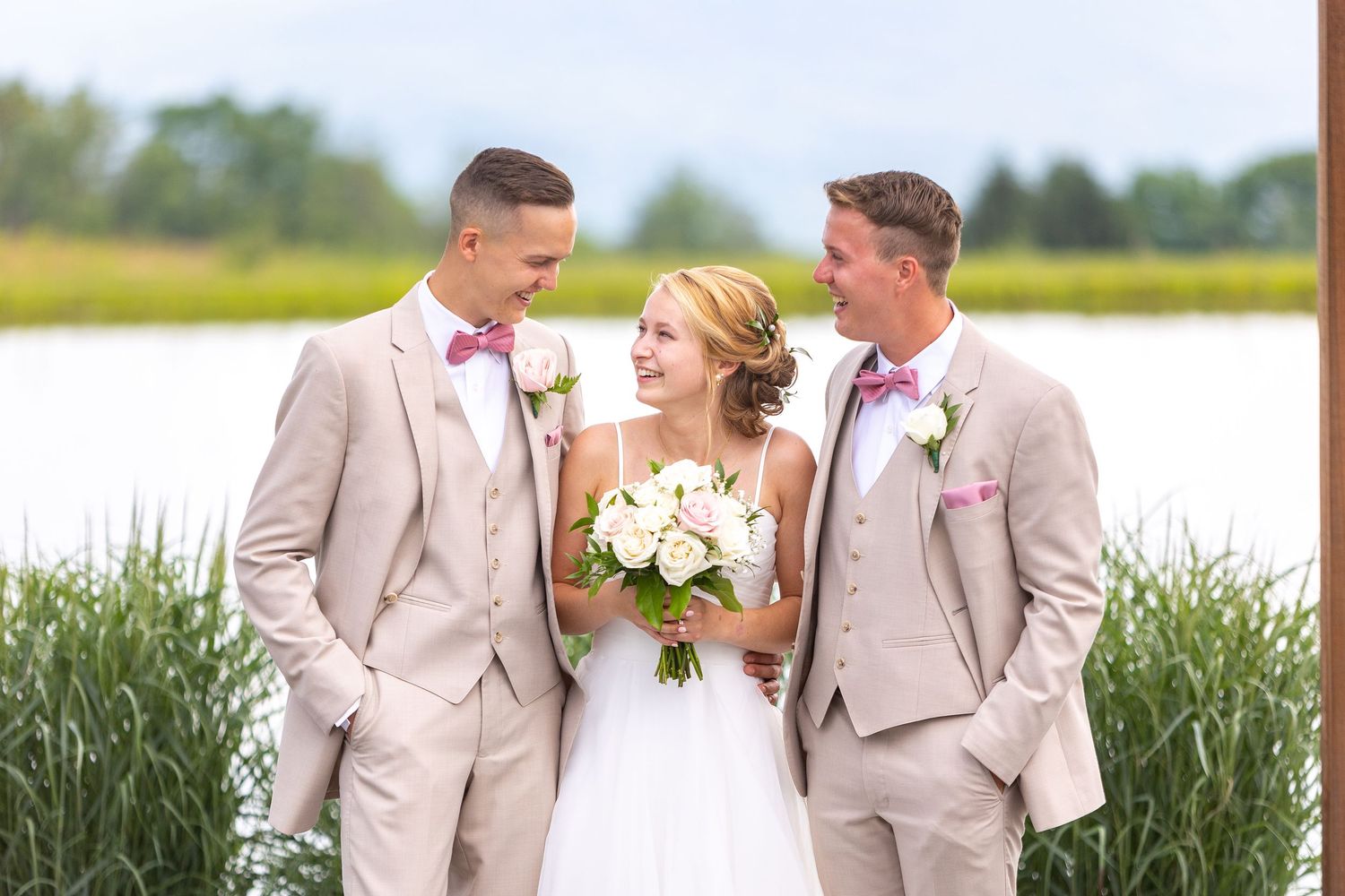 Wedding couple and wedding party in beige suits share joyful moments by a scenic waterfront on their special day.