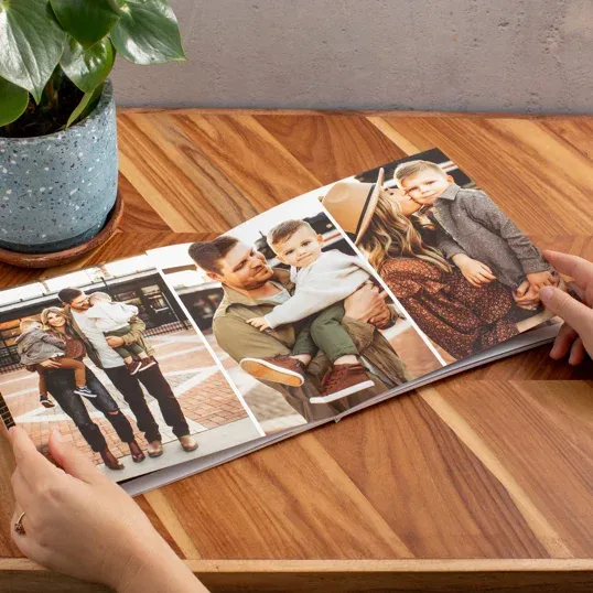 Someone flips through a photo album containing printed pictures on a wooden table with a potted plant nearby.