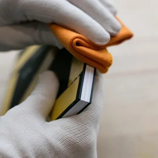 White-gloved hands wiping down a book spine with an orange cloth for maintenance and preservation.