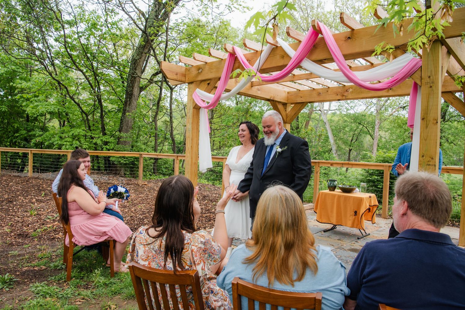 Casual outdoor wedding ceremony with guests seated on wooden chairs beneath a decorated pergola.