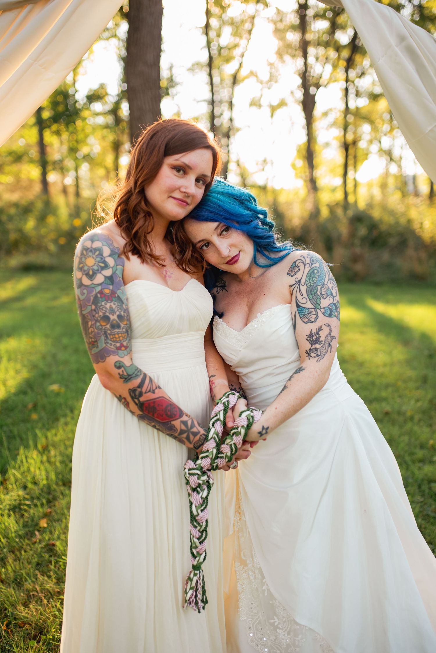 Two brides in white wedding dresses showing off their colorful tattoo sleeves at sunset.