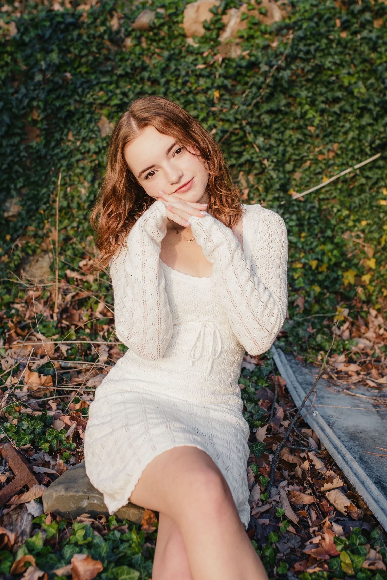 Woman in white dress posing on wooden bench surrounded by green ivy.