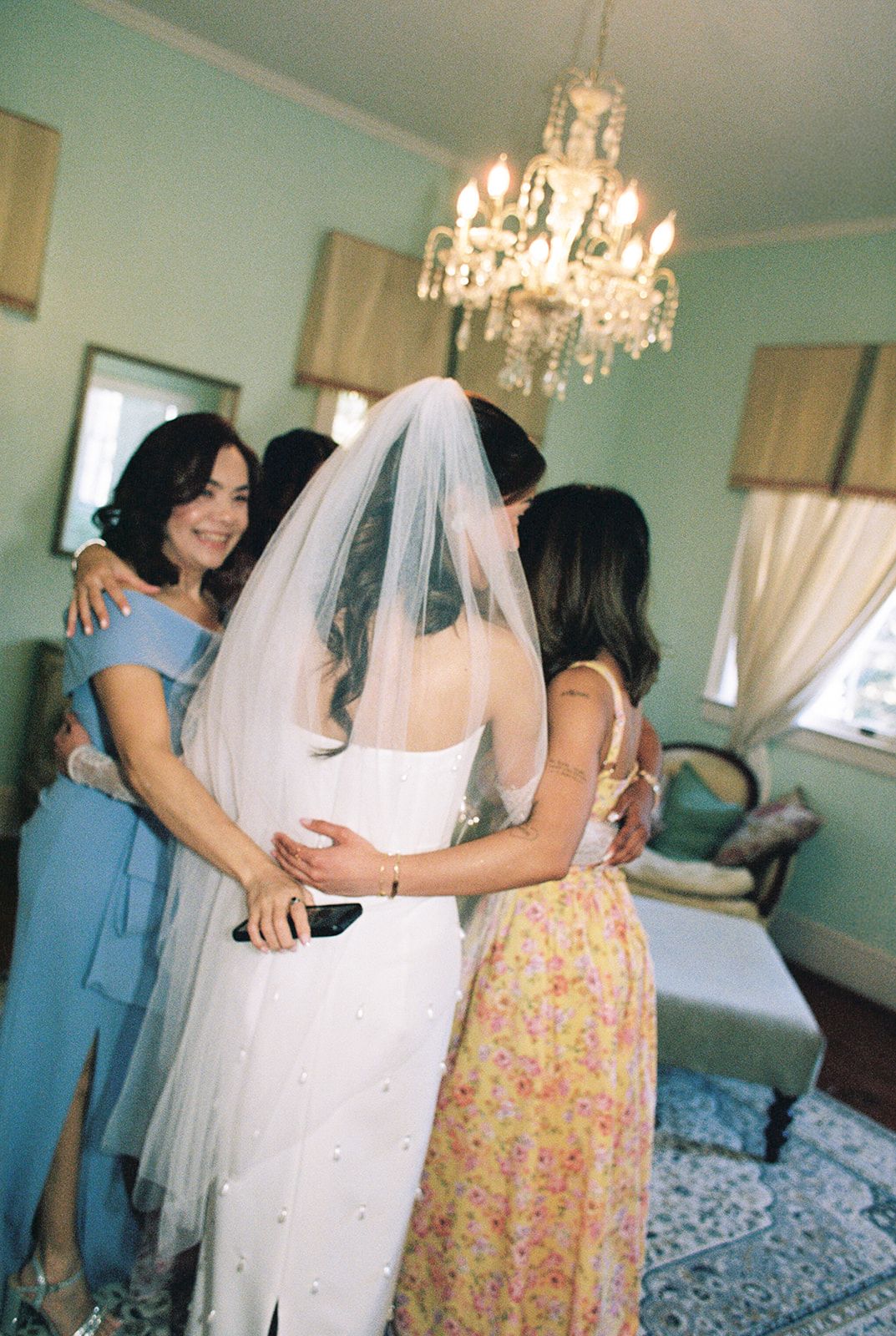 Bridesmaids in pastel dresses embrace a bride in white under an elegant crystal chandelier in a mint-colored room.