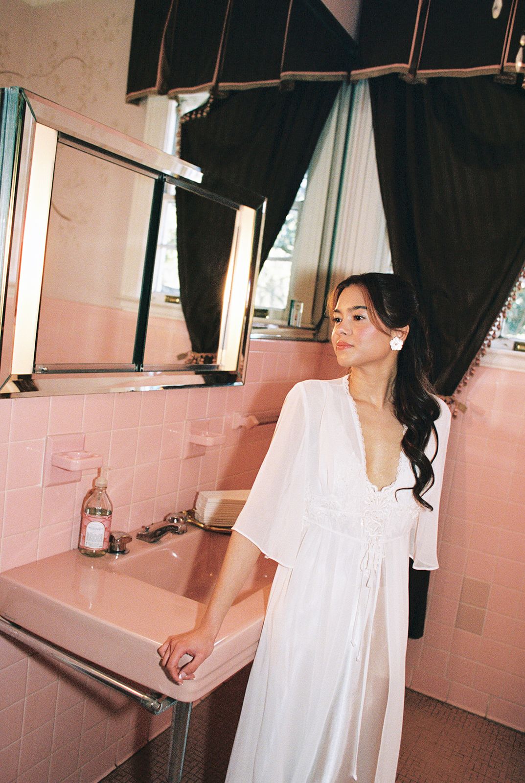 Figure in flowing white dress stands in retro pink-tiled bathroom with dark curtains and vintage decor.