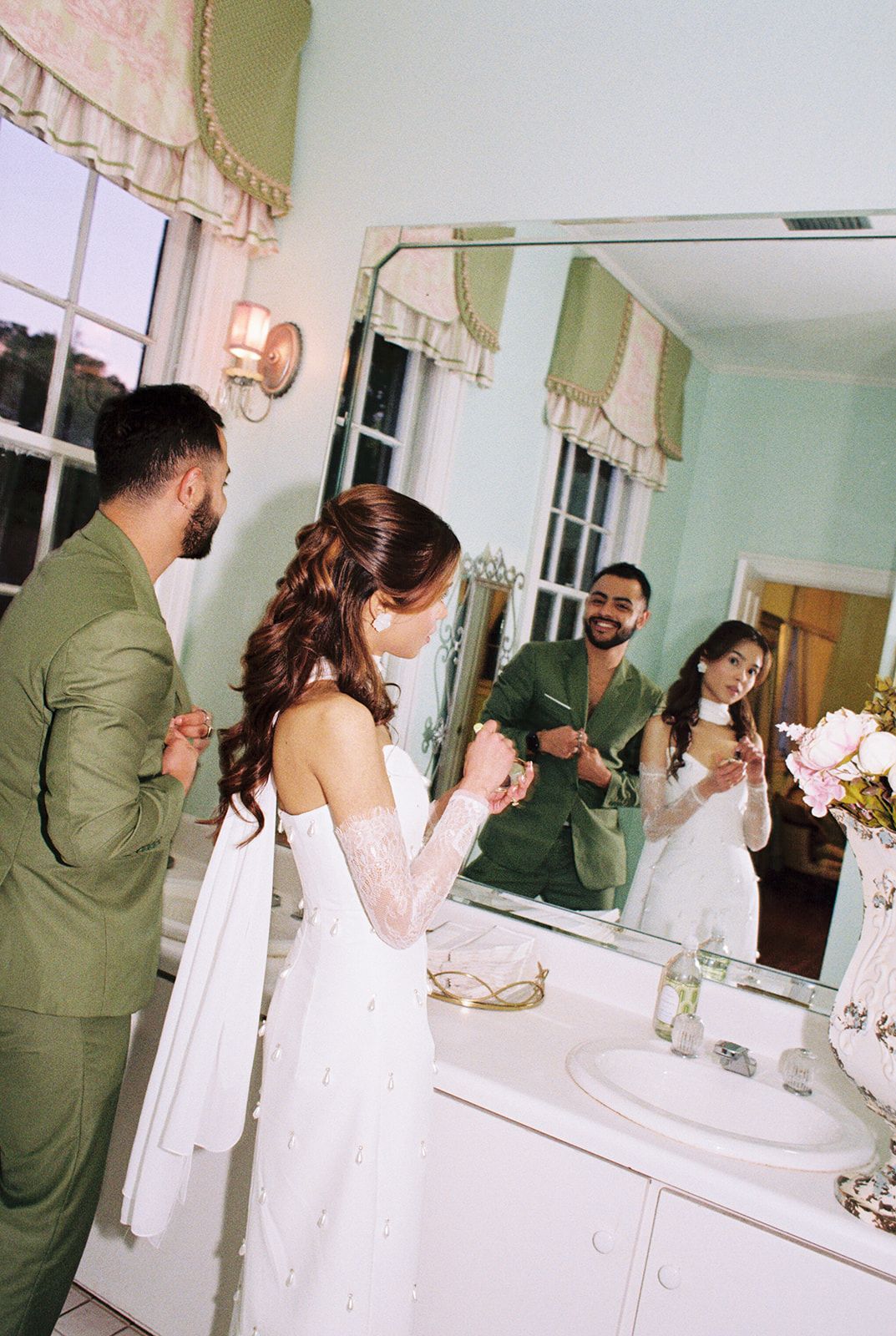 Couple stands at bathroom vanity mirror with matching green and white outfits during special occasion.