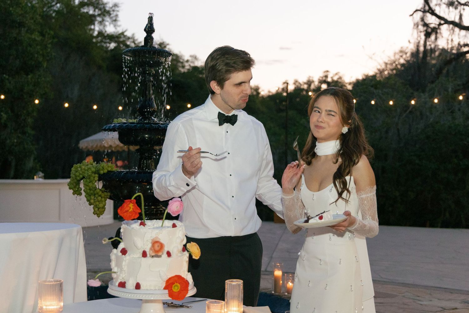 Two figures share a slice of wedding cake in front of string lights at an outdoor evening reception.