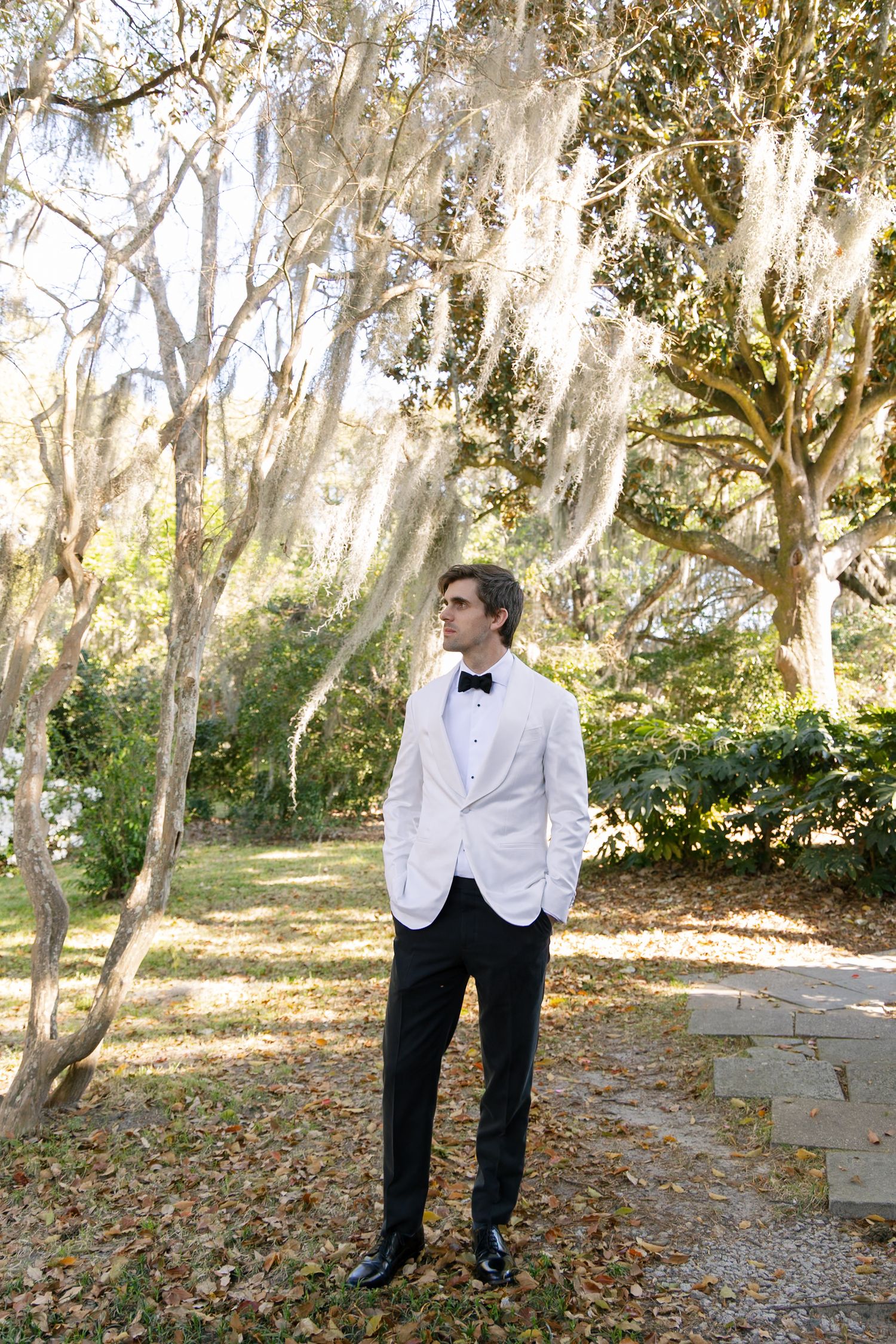 Person in a formal black and white tuxedo stands thoughtfully among tall trees in a natural outdoor setting.