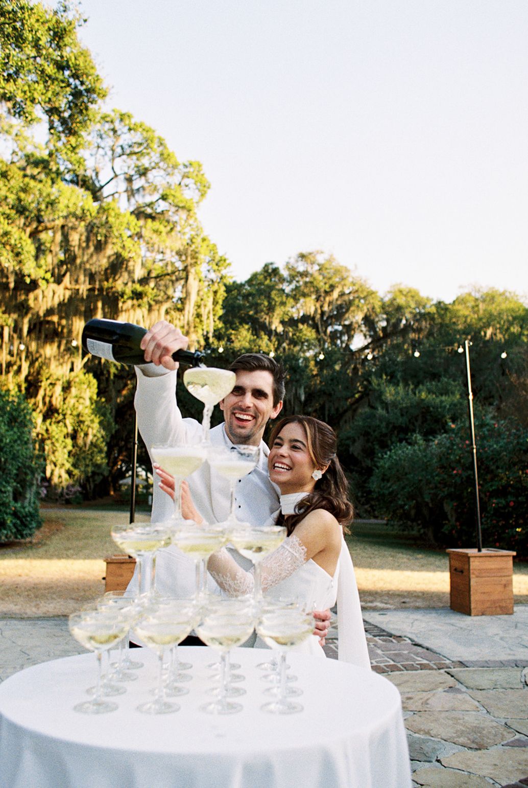 Couple pouring champagne into tower of glasses at outdoor celebration surrounded by Spanish moss trees at dusk.