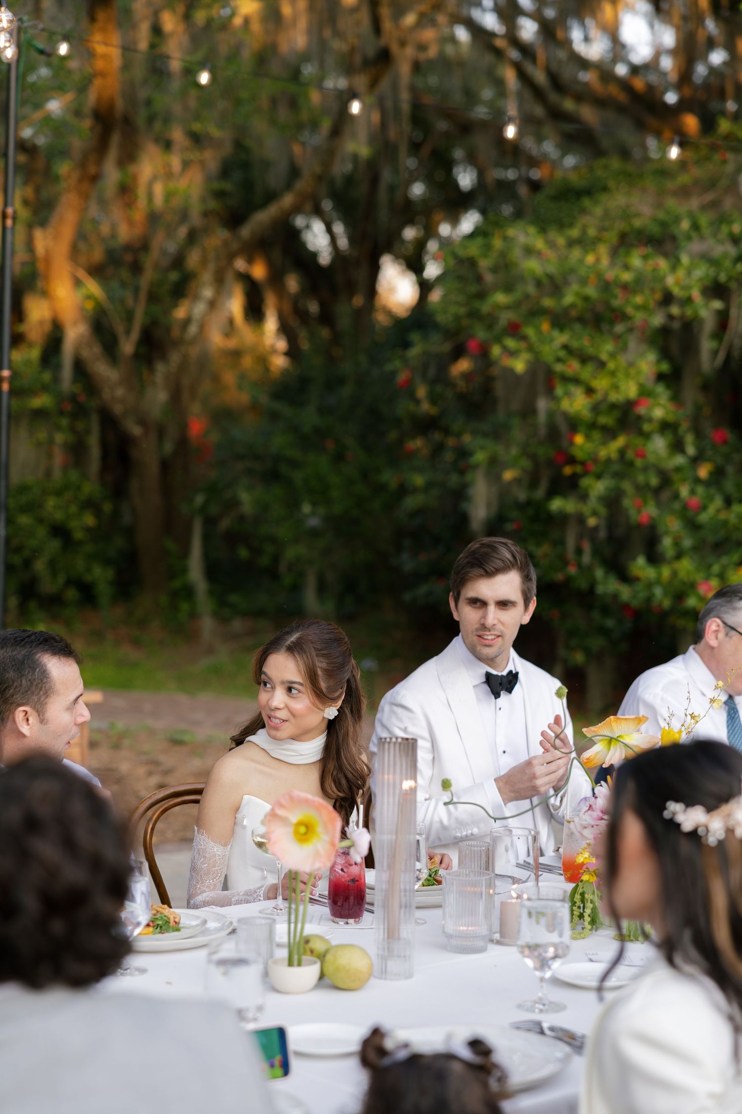 Guests dining outdoors at an elegant wedding reception with white linens and floral centerpieces under moss-draped trees.