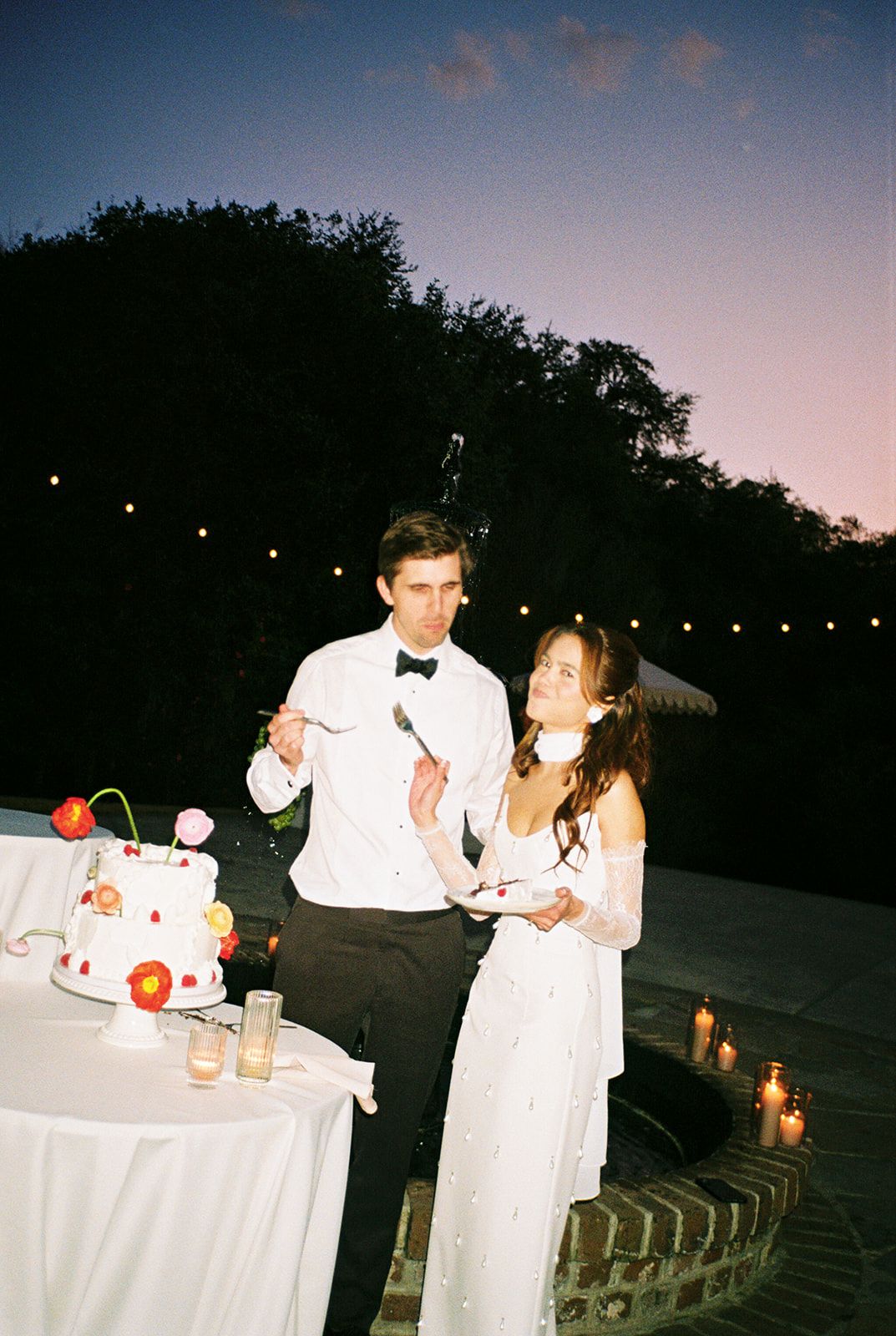 A couple shares a dessert moment at an outdoor evening reception with string lights and candles illuminating the scene.