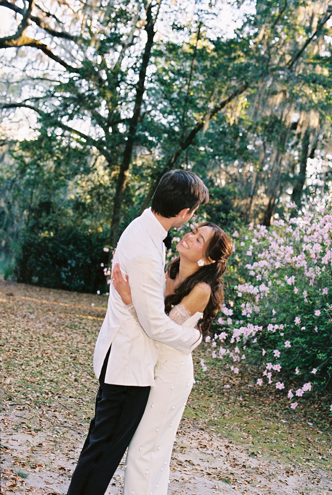 A romantic couple kisses in a dreamy outdoor garden setting with blooming pink flowers and trees in the background.