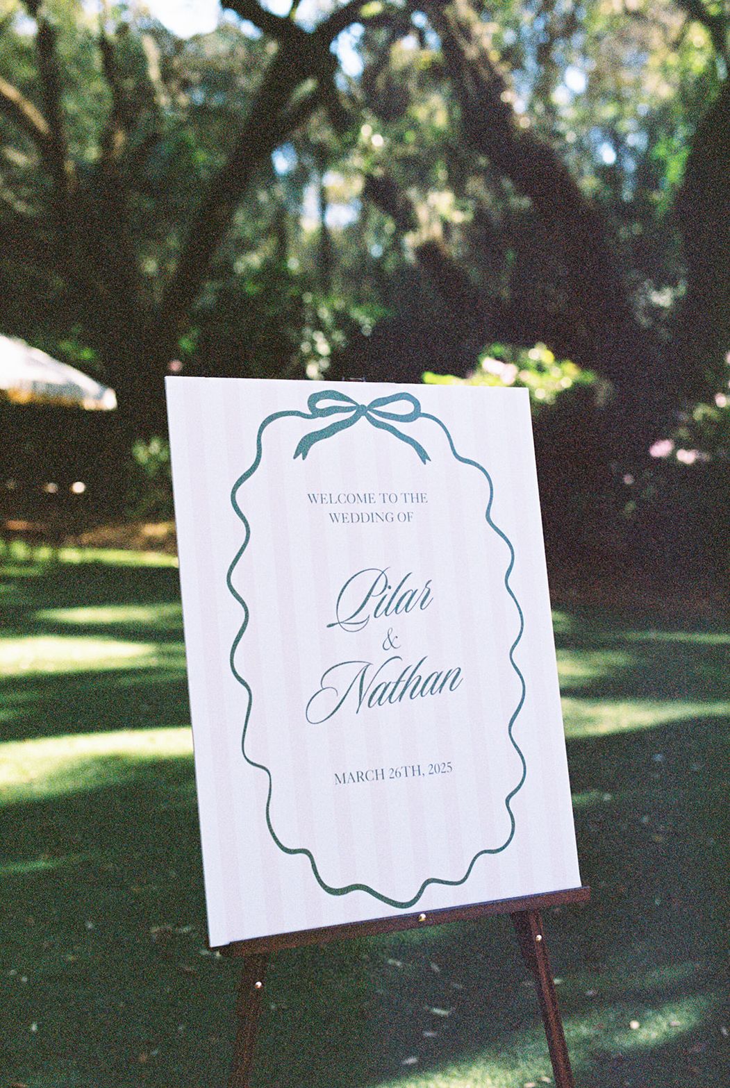 Elegant white wedding signs with green text displaying couple's names and date mounted on posts in a sunny garden setting.