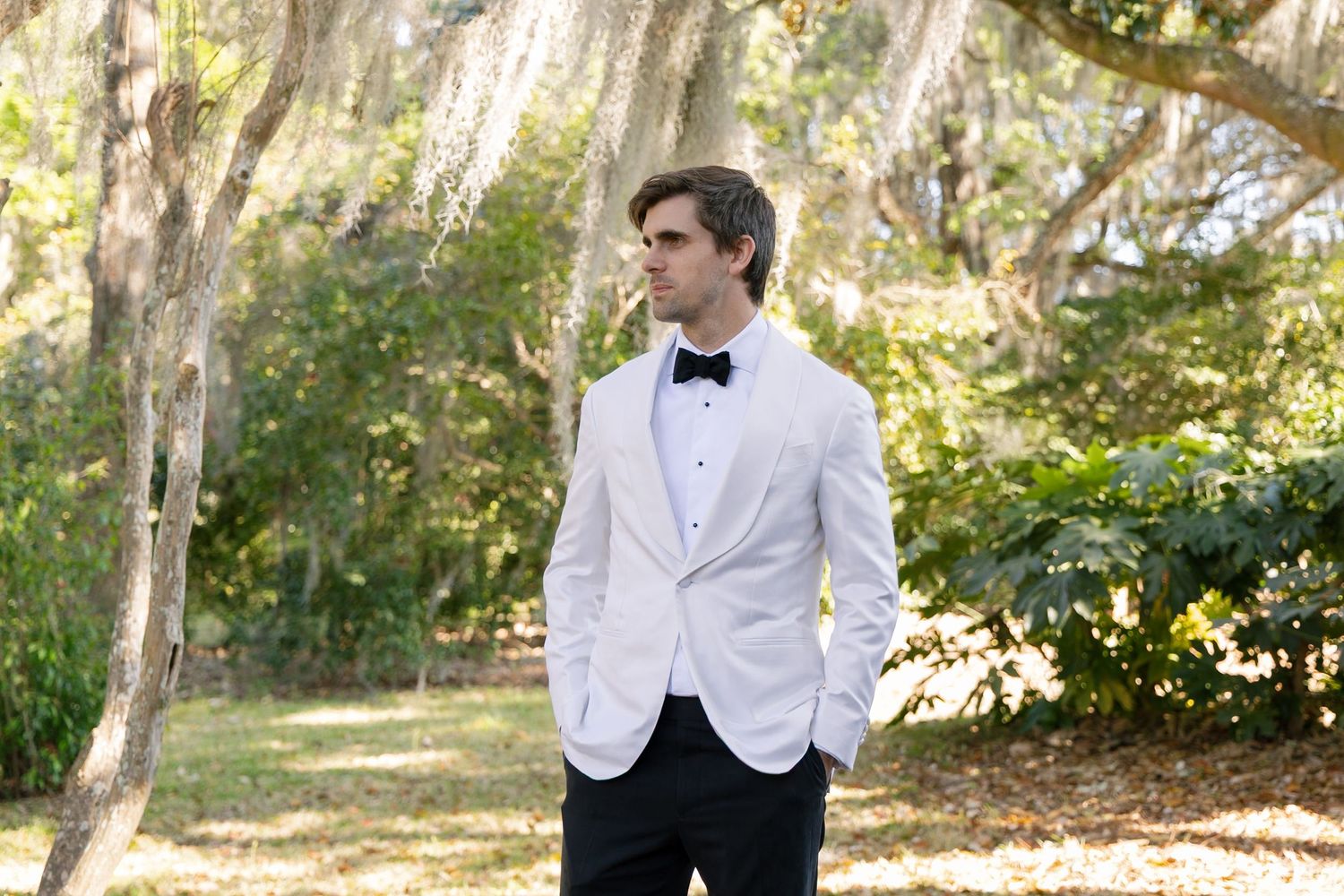 Figure in crisp white dinner jacket and bow tie poses outdoors in dappled sunlight among trees.