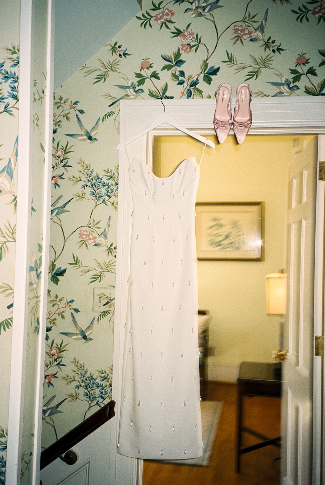 A white beaded wedding dress hangs on a doorway with pink heels on top, against floral wallpaper with green and pink details.