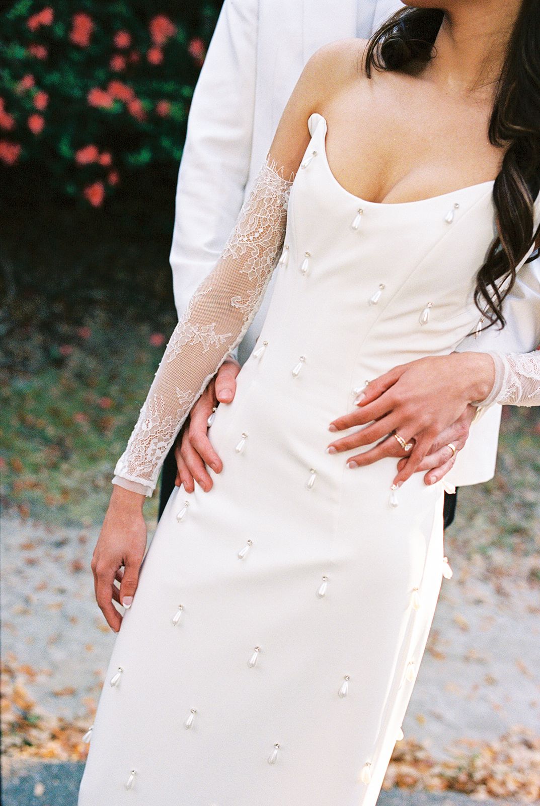 Close-up view of elegant white wedding dress with sheer lace sleeve and delicate floral appliques against autumn foliage.